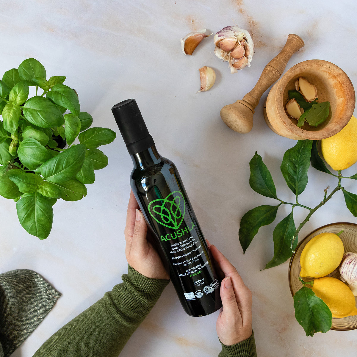 Holding a 500 ml Acushla Organic DOP Trás-os-Montes bottle near basil, lemons, and garlic.