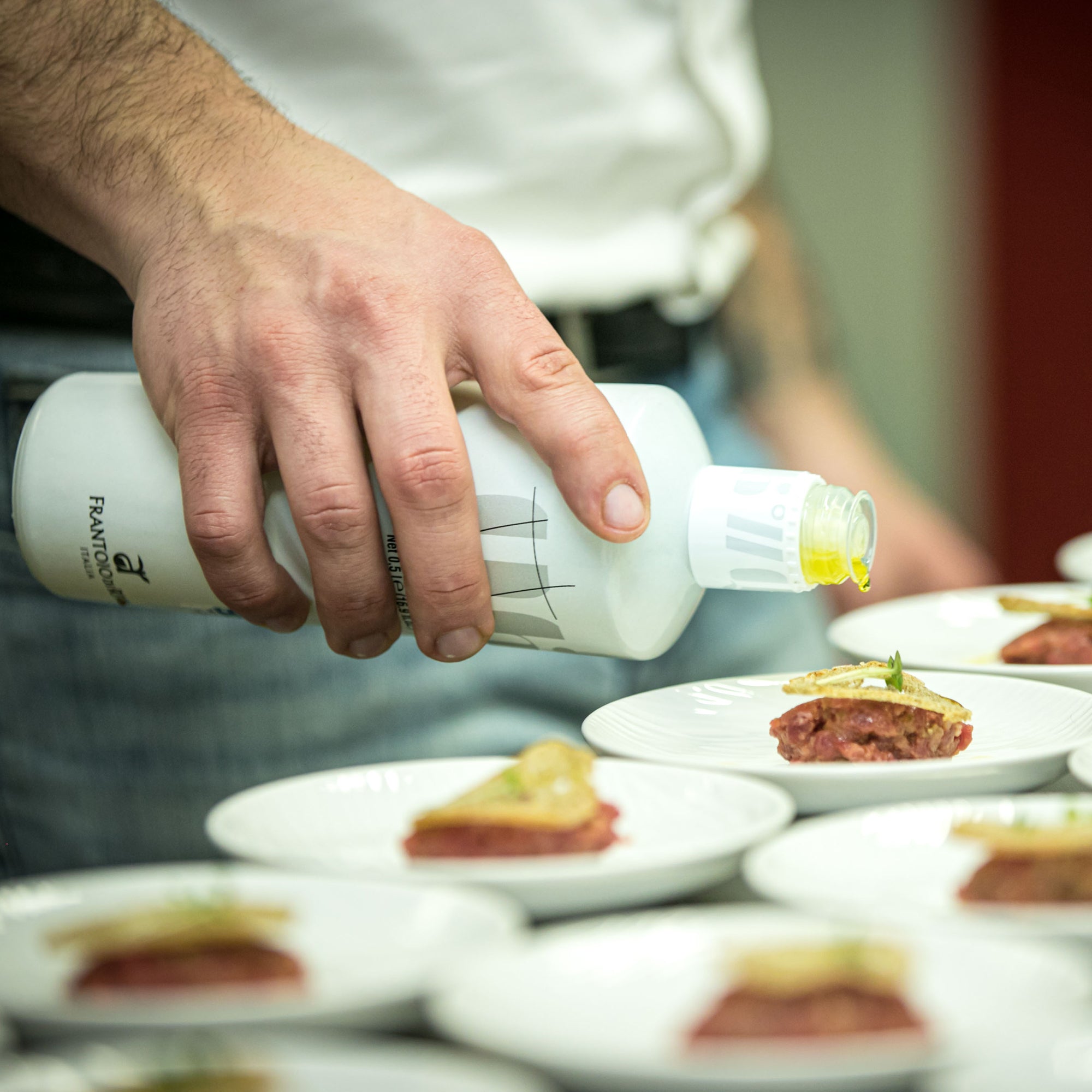 Chef pouring 46° Parallelo Organic 500 ml olive oil over plated food during preparation.