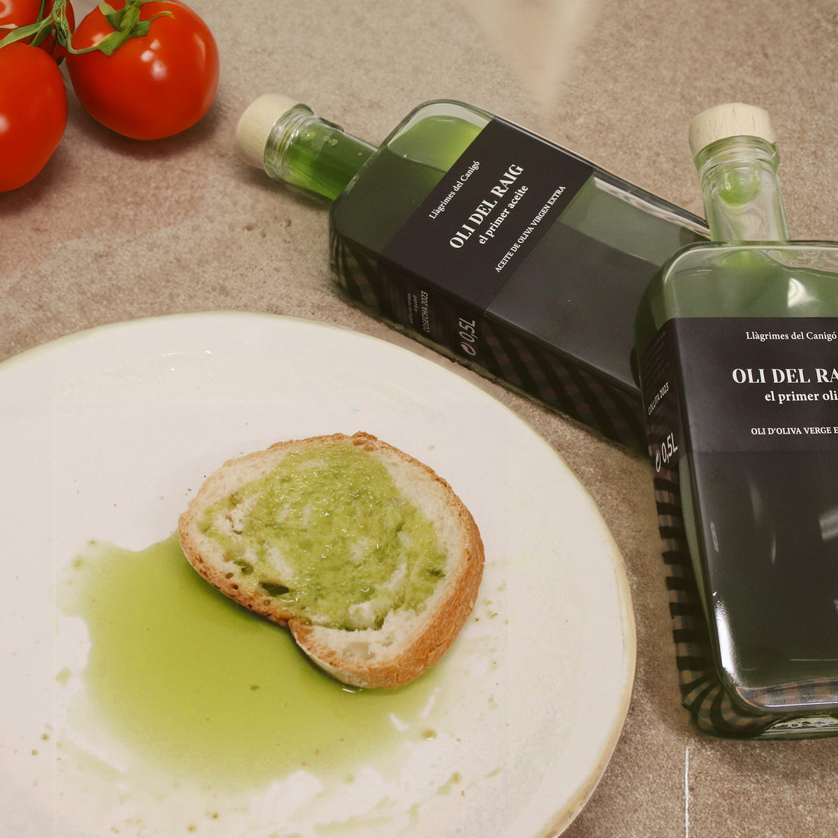 Two 500 ml Oli del Raig Novello bottles beside tomatoes and bread soaked with green olive oil.