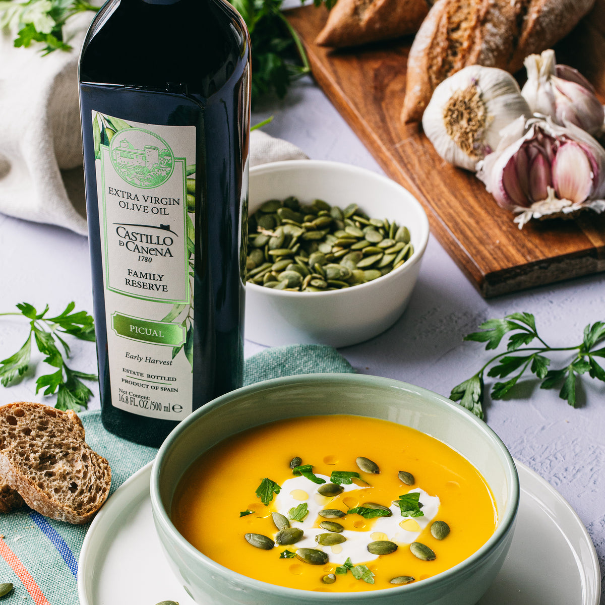 500 ml bottle of Castillo de Canena Family Reserve Picual beside pumpkin soup, bread, garlic, parsley, and pumpkin seeds.