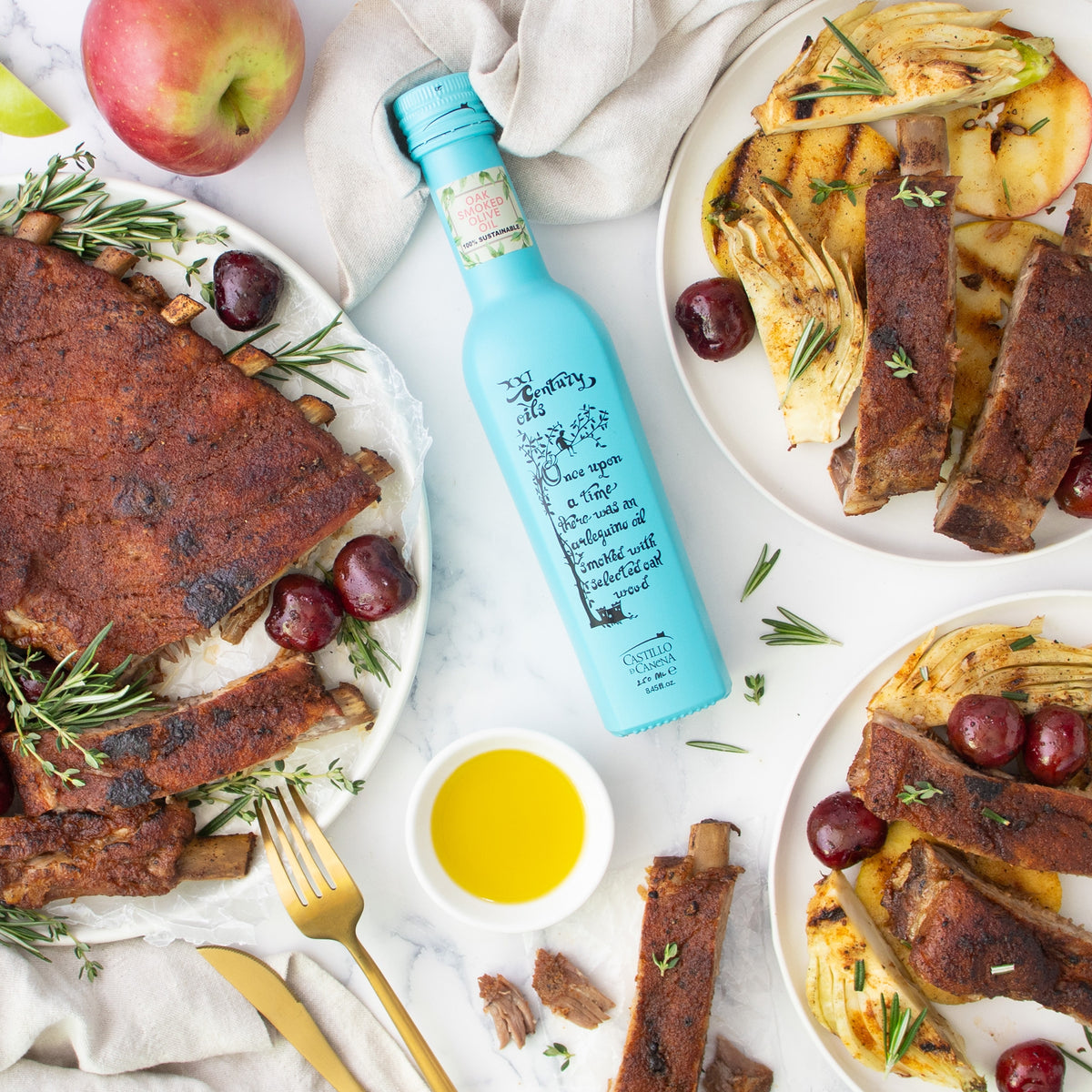 Castillo de Canena Smoked Arbequina bottle surrounded by plates of roasted ribs, apples, fennel, grapes, and herbs.