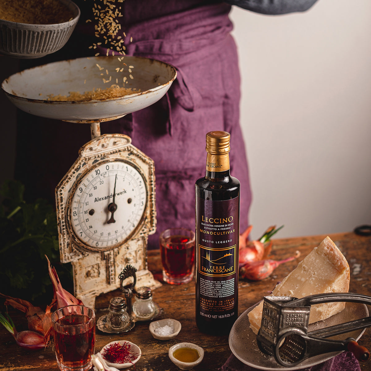 Terre Francescane Leccino 500 ml bottle beside a vintage kitchen scale, Parmesan, seasonings, and cooking utensils.