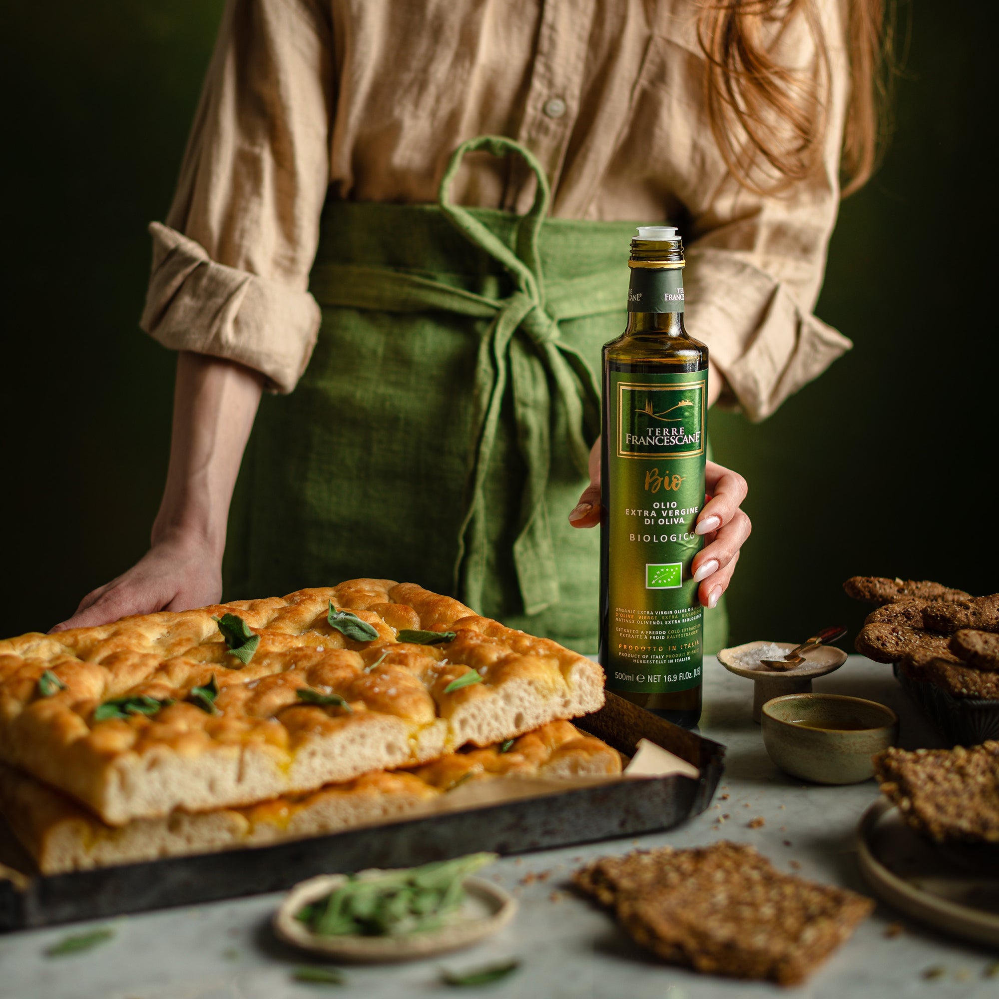 Holding Terre Francescane Organic 500 ml olive oil bottle beside freshly baked focaccia, crackers, and breadsticks.