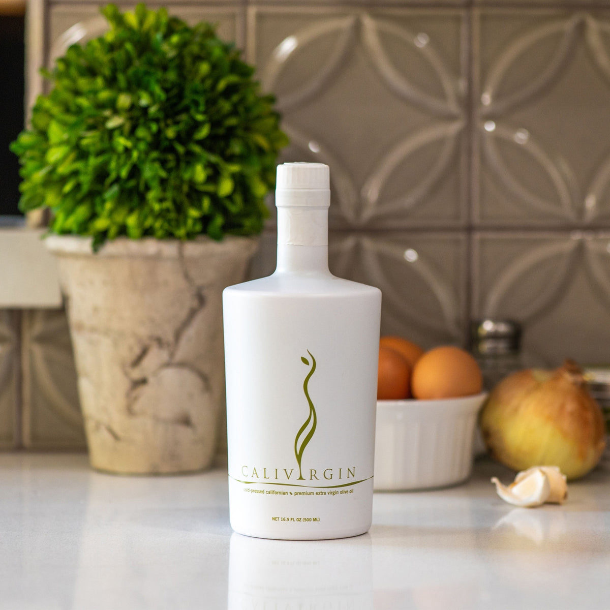 Calivirgin Premium 500 ml bottle on a kitchen counter with herbs, citrus, onion, and garlic.