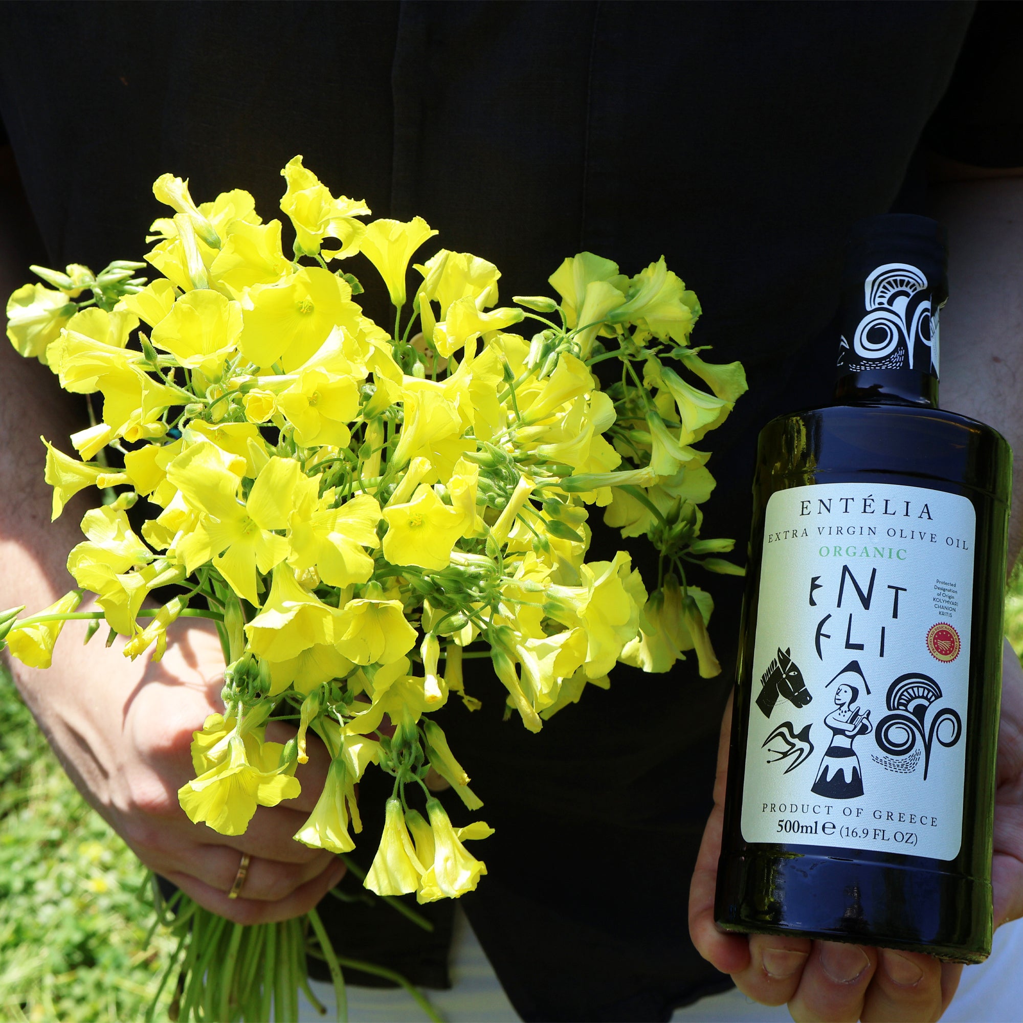 A person holding yellow flowers and a 500 ml bottle of Entelia Organic extra virgin olive oil.