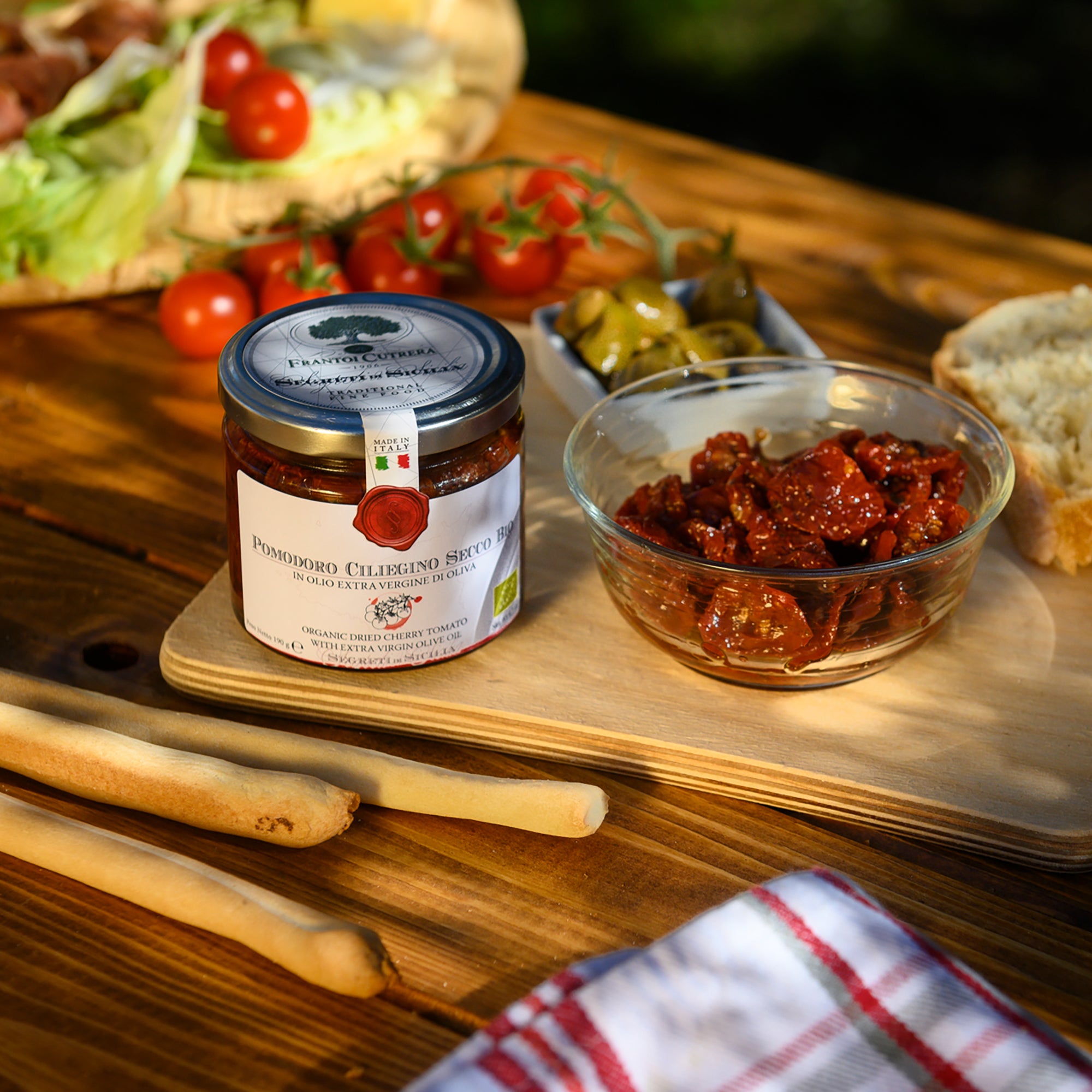 Frantoi Cutrera Organic Dried Cherry Tomatoes jar beside a serving bowl with bread and fresh tomatoes.