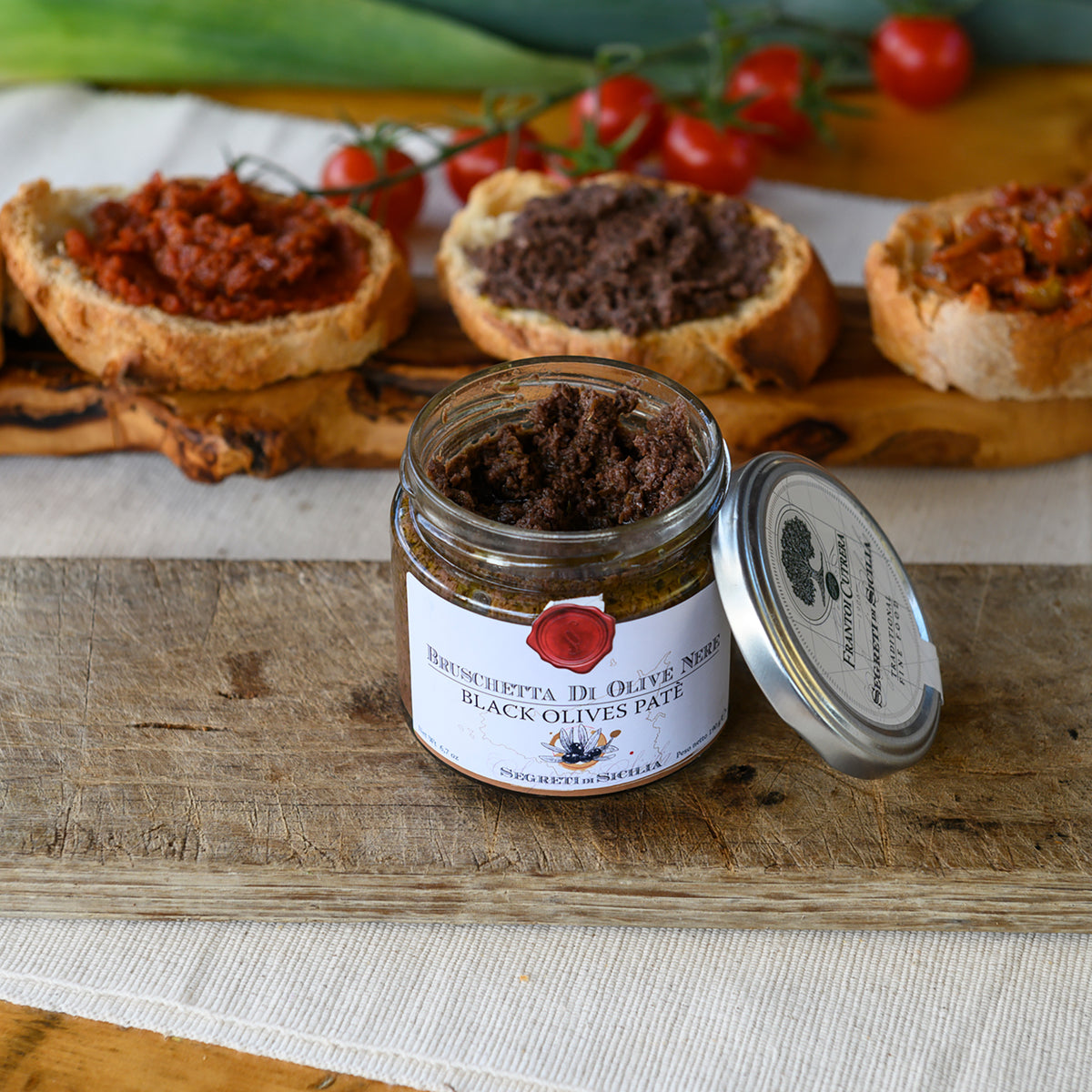 Open jar of Frantoi Cutrera Tonda Iblea Black Olive Pate beside rustic bread slices.