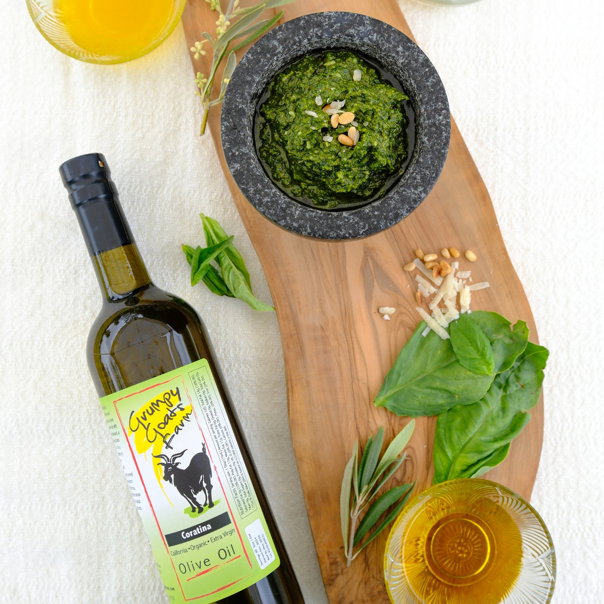 Grumpy Goats Organic Coratina olive oil bottle beside fresh basil, pesto, pine nuts, and cheese.