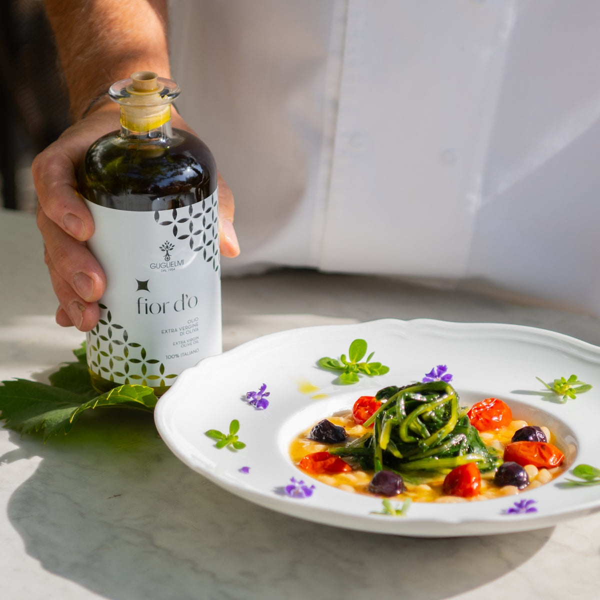 Chef holds Guglielmi Fior D’O Nuovo olive oil beside a vegetable dish.