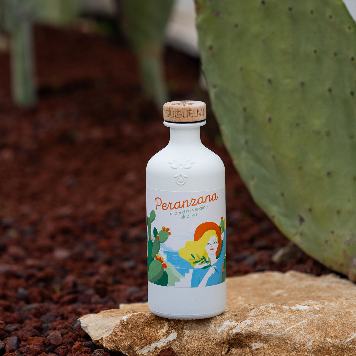 Guglielmi Peranzana 500 ml extra virgin olive oil bottle on a rock beside large prickly pear cactus pads.