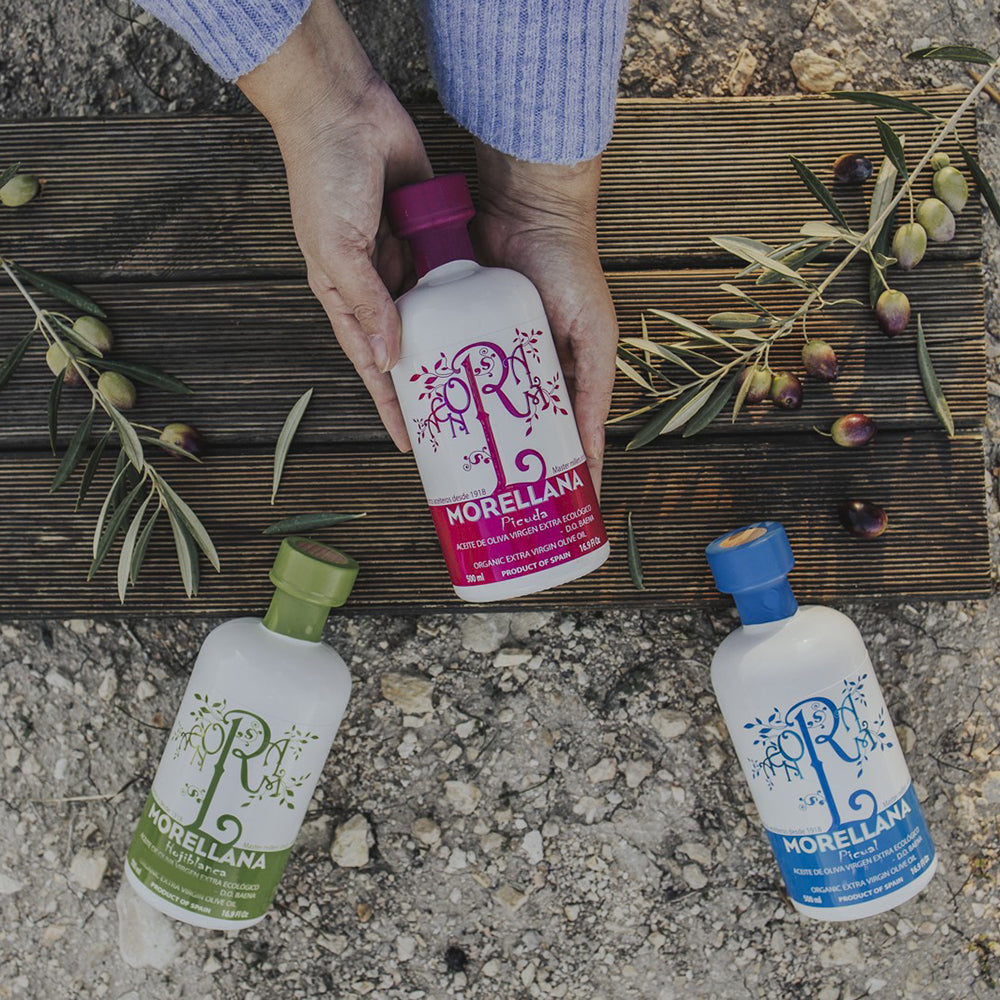Holding Morellana Organic Picuda among Morellana Picual and Hojiblanca bottles, olive branches, and freshly harvested olives.