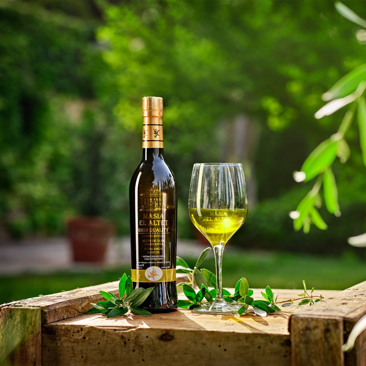 Masia el Altet High Quality 500 ml bottle beside a glass of olive oil in a sunlit garden.