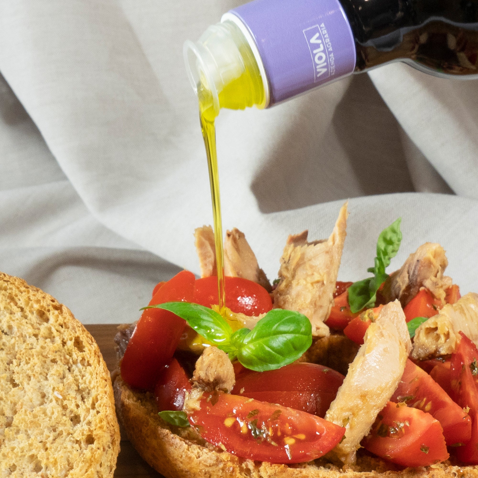 Viola extra virgin olive oil being poured over a bread with tomatoes, tuna, and basil.