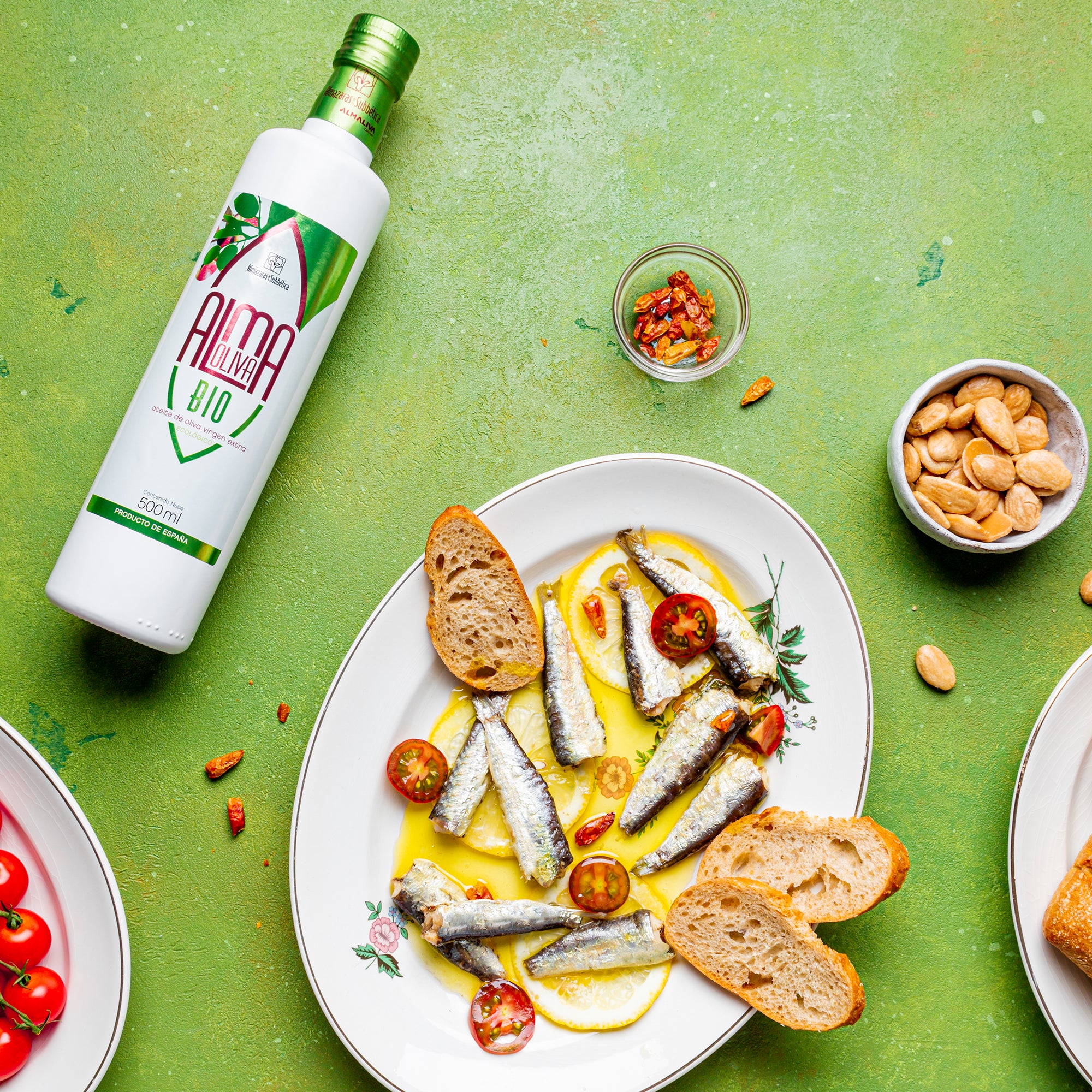 Almaoliva Bio 500 ml bottle beside sardines in olive oil, bread, tomatoes, almonds, and dried peppers.