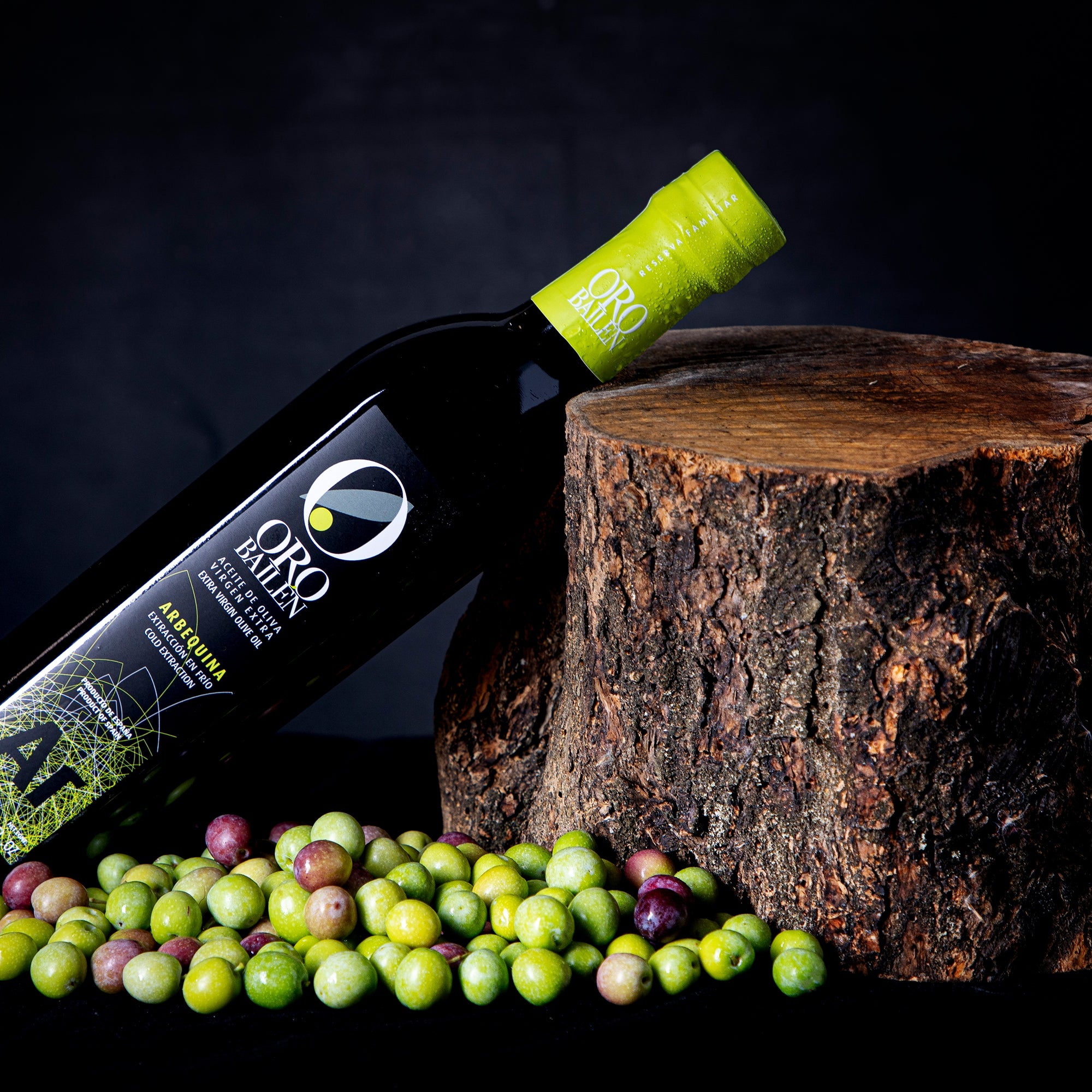 Oro Bailen Reserva Familiar Arbequina bottle leaning beside a tree stump and scattered olives.