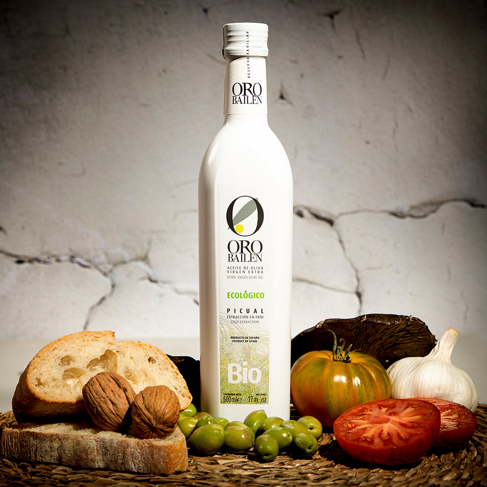 Oro Bailen Picual Organic bottle beside bread, walnuts, olives, tomatoes, garlic, and mushrooms.