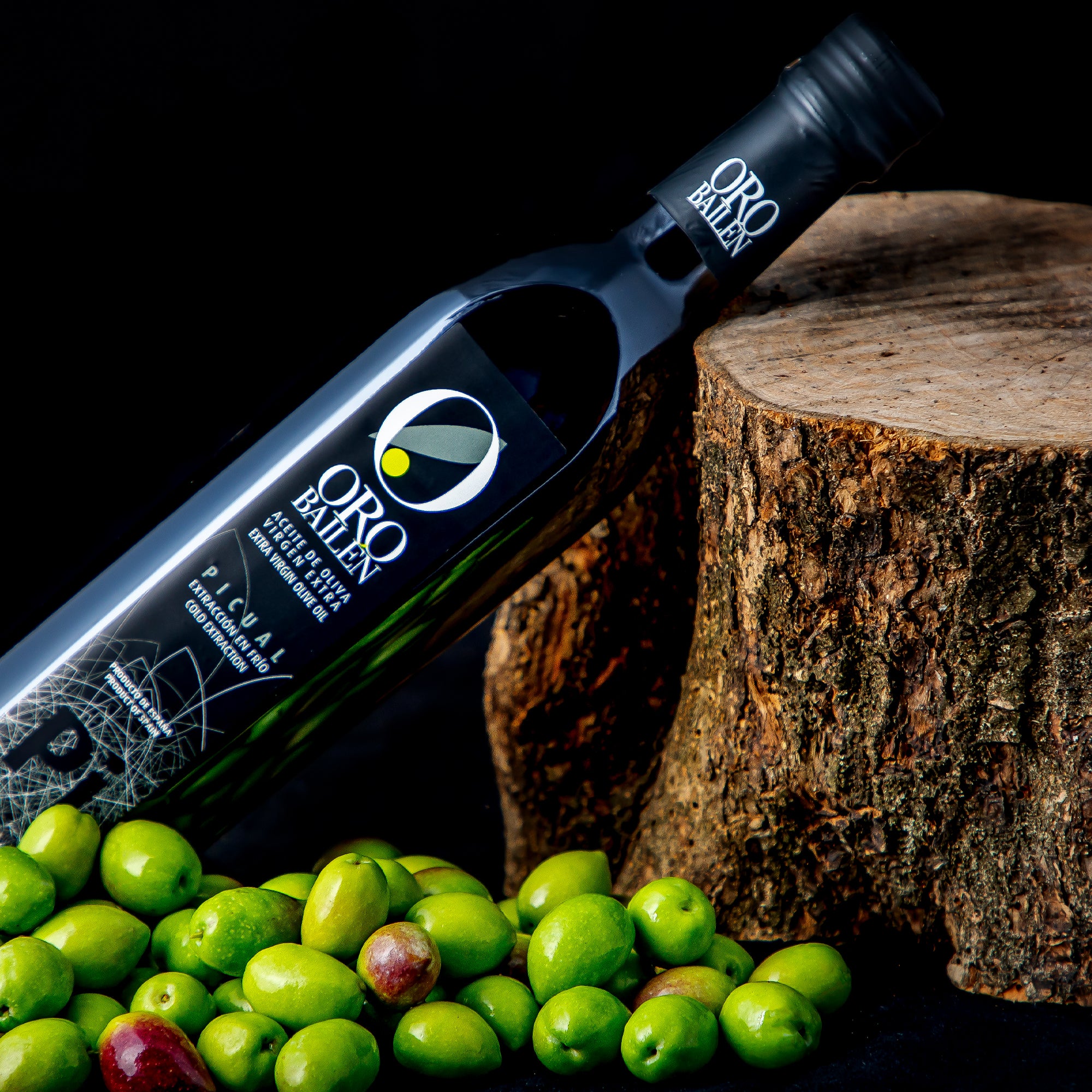 Oro Bailen Reserva Familiar Picual bottle beside a log and scattered fresh olives.