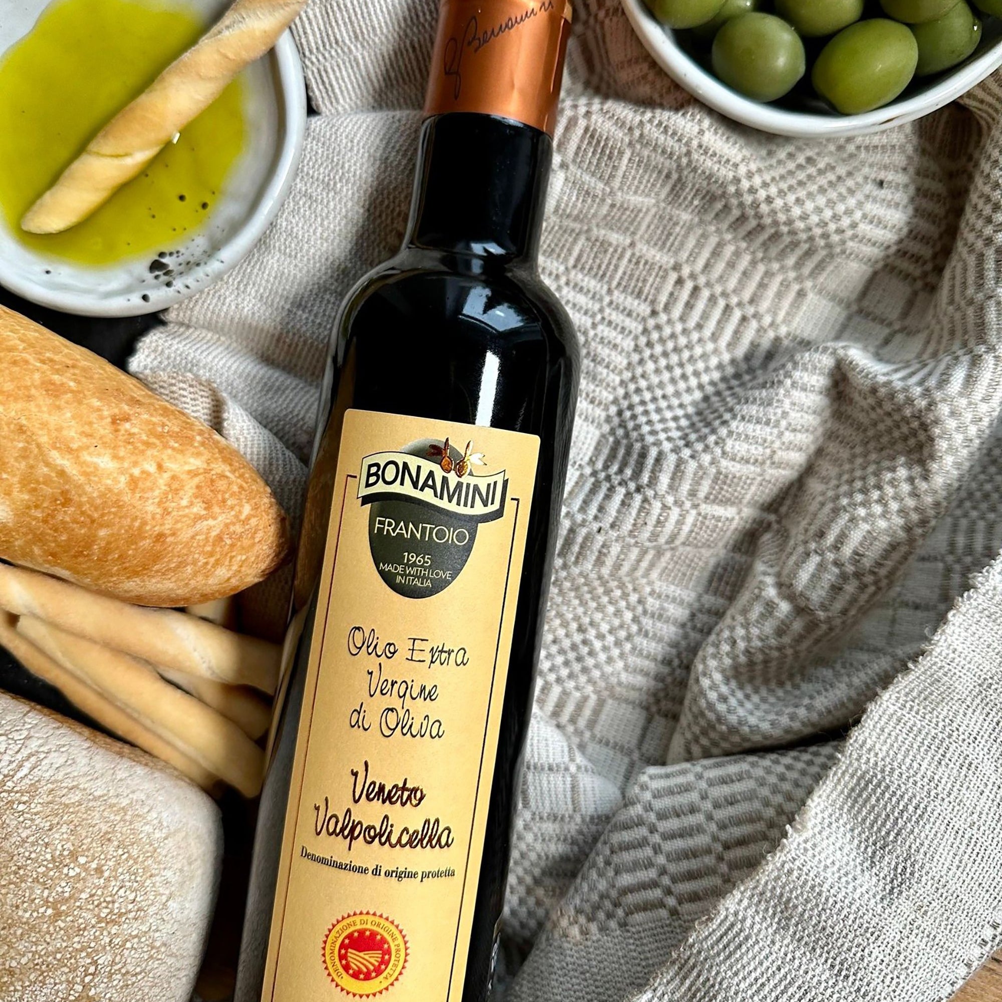 Frantoio Bonamini DOP Veneto Valpolicella bottle beside bread, breadsticks, and green olives.