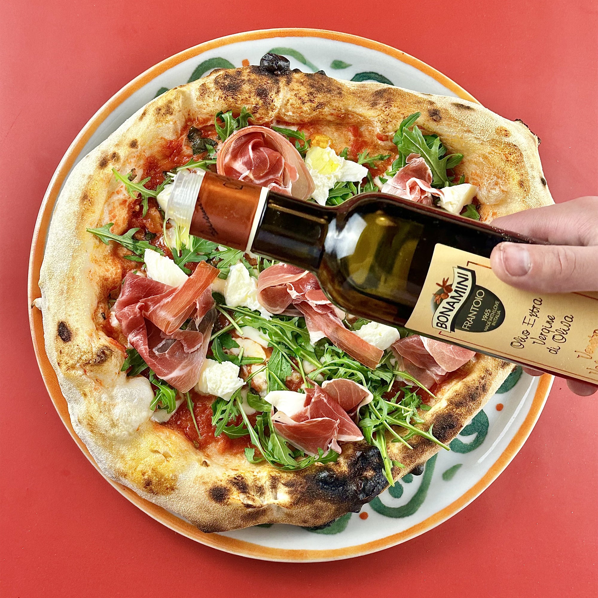 Frantoio Bonamini olive oil being poured over a pizza topped with prosciutto, arugula, and cheese.