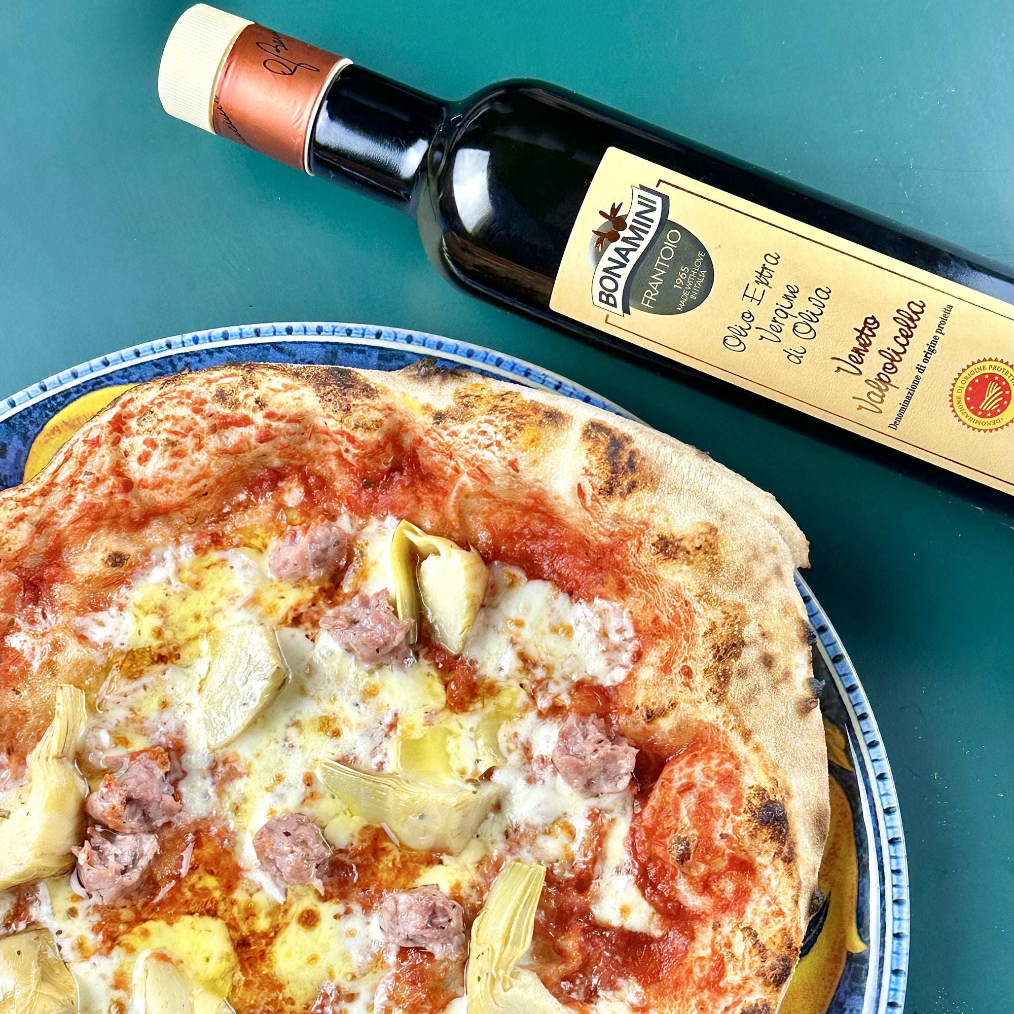 Frantoio Bonamini DOP Veneto Valpolicella bottle beside a pizza topped with artichokes, sausage, and cheese.