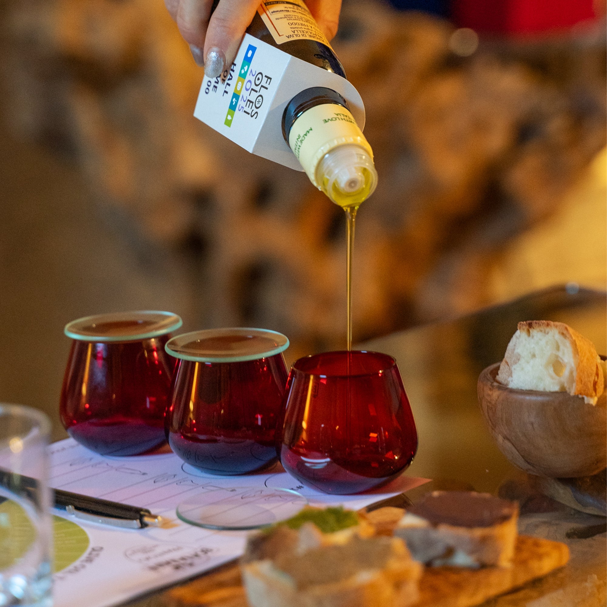 Olive oil poured from a Frantoio Bonamini bottle into a red tasting glass.