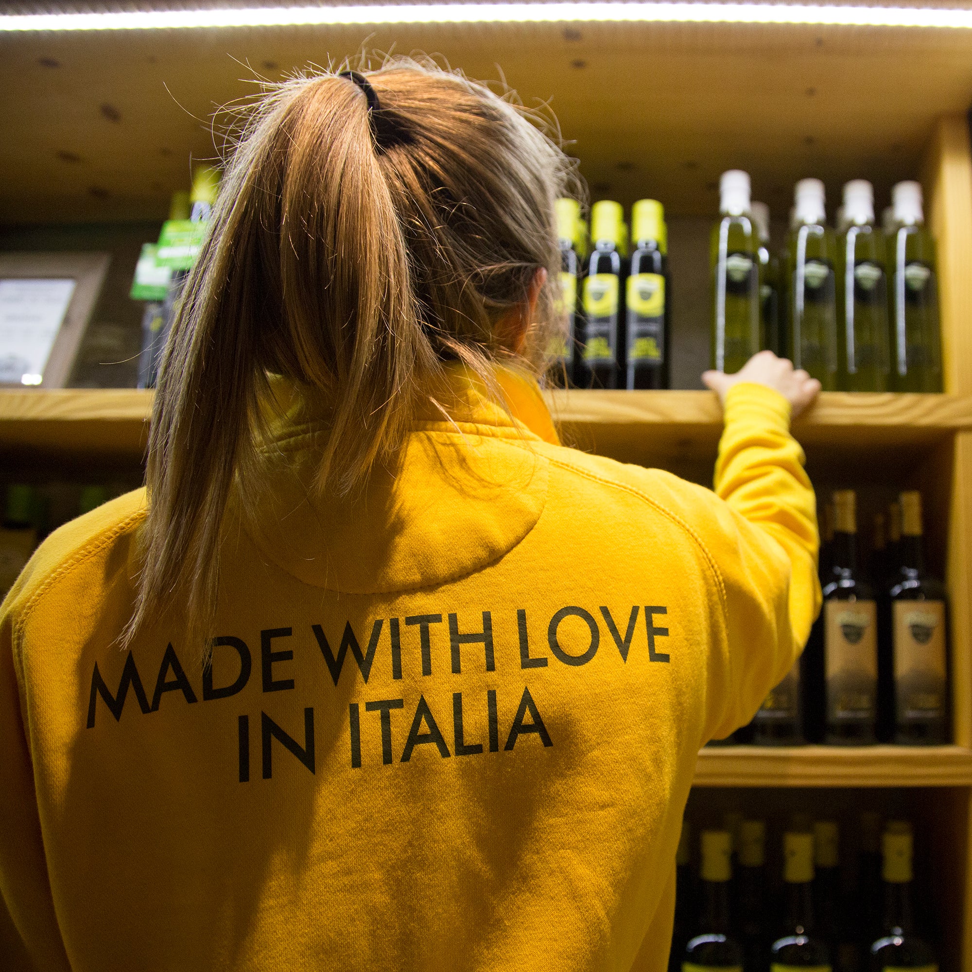 A woman arranges Bonamini bottles on shelves, wearing a yellow “Made With Love in Italia” sweatshirt.