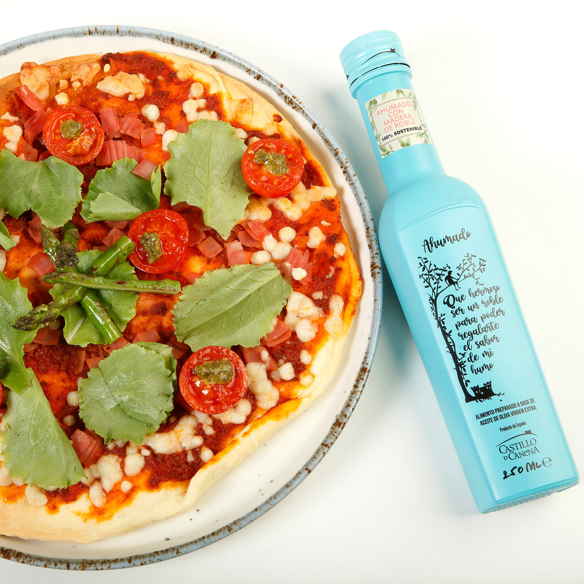 A 250 ml bottle of Castillo de Canena Smoked Arbequina beside a pizza topped with tomatoes, asparagus, cheese, and leafy greens.