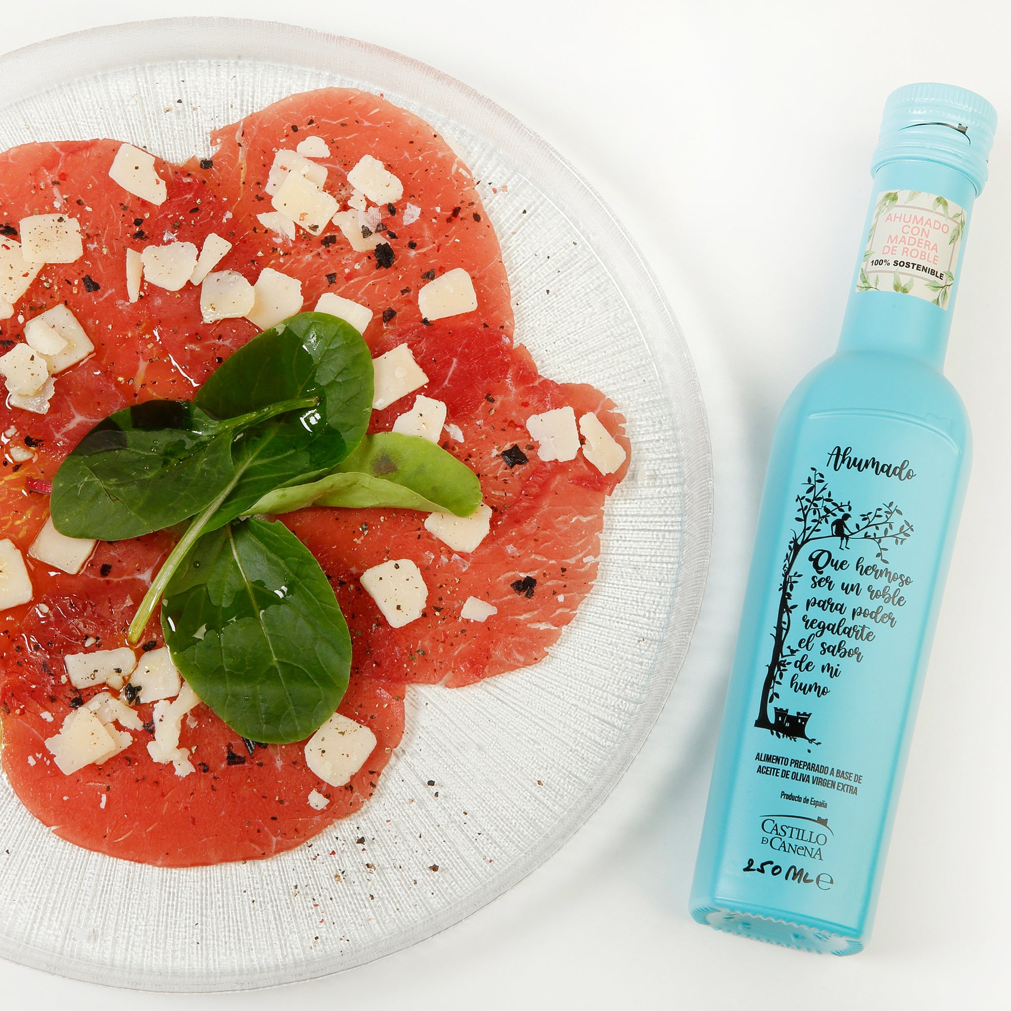 250 ml bottle of Castillo de Canena Smoked Arbequina bottle beside beef carpaccio topped with cheese, spinach, olive oil, and pepper.