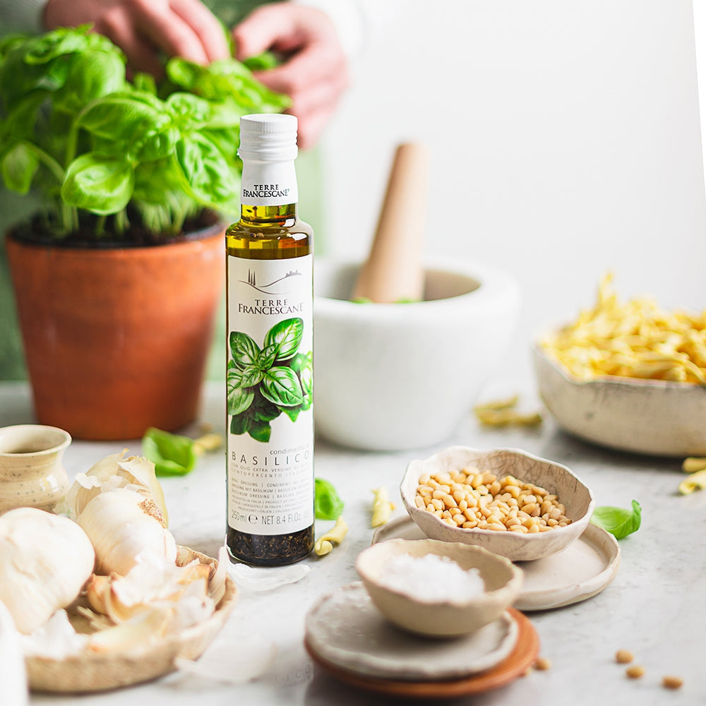 Terre Francescane Basil 250 ml infused extra virgin olive oil beside fresh basil, garlic, pine nuts, and pasta.