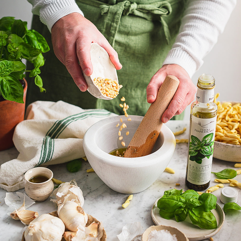 Person adds pine nuts to a mortar beside Terre Francescane Basil 250 ml olive oil and fresh basil.