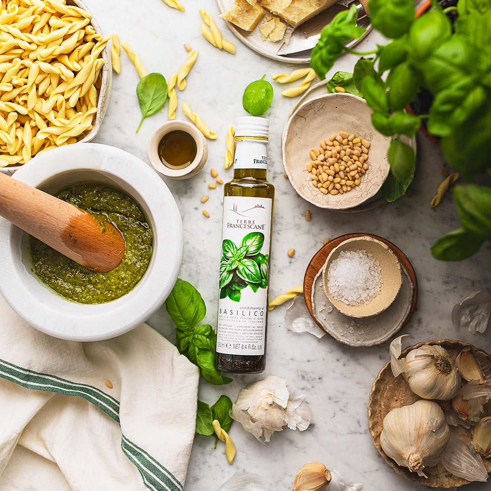 Terre Francescane Basil 250 ml infused extra virgin olive oil beside pesto ingredients, pasta, basil, garlic, and nuts.