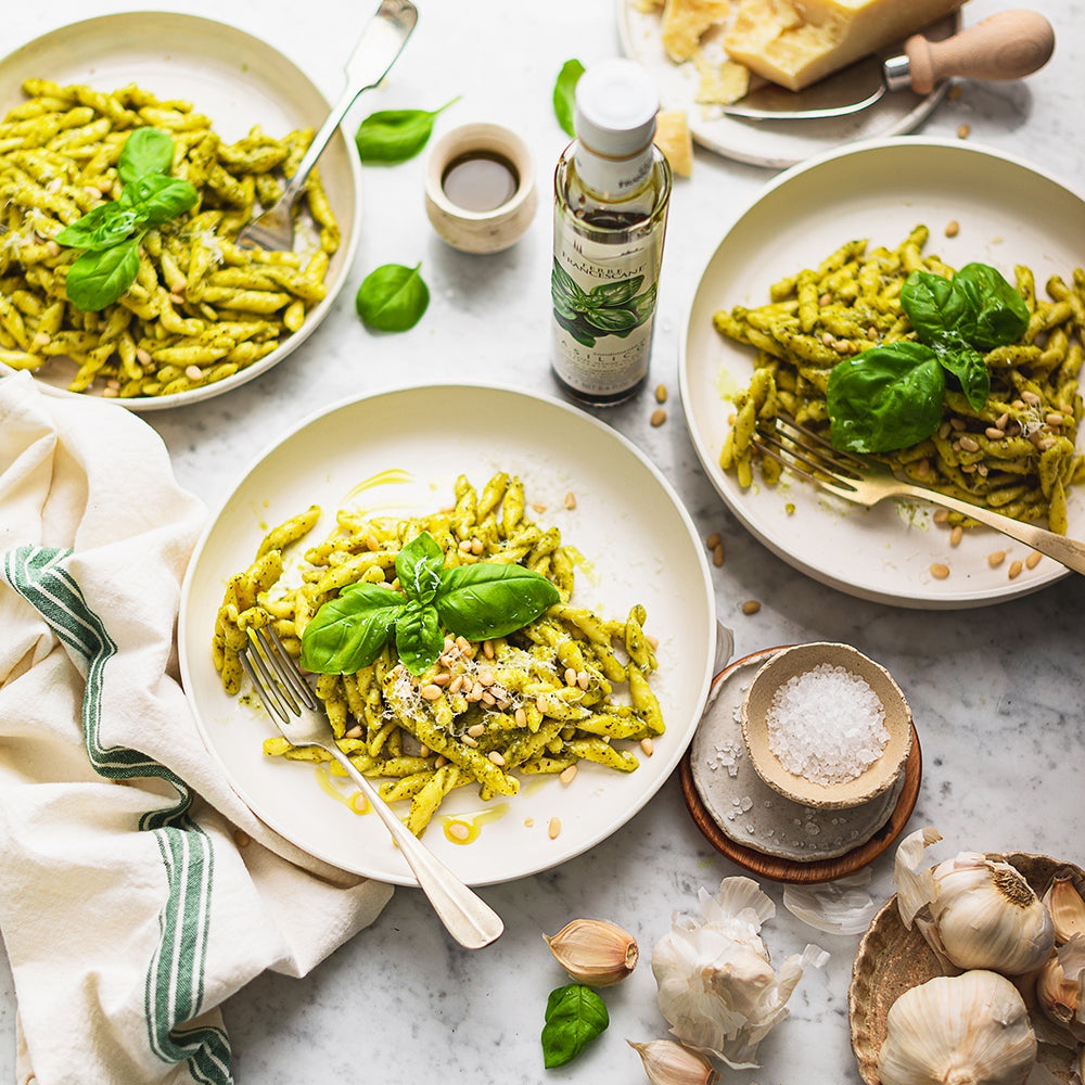 Plates of pesto pasta with basil and pine nuts and a Terre Francescane Basil olive oil in between.
