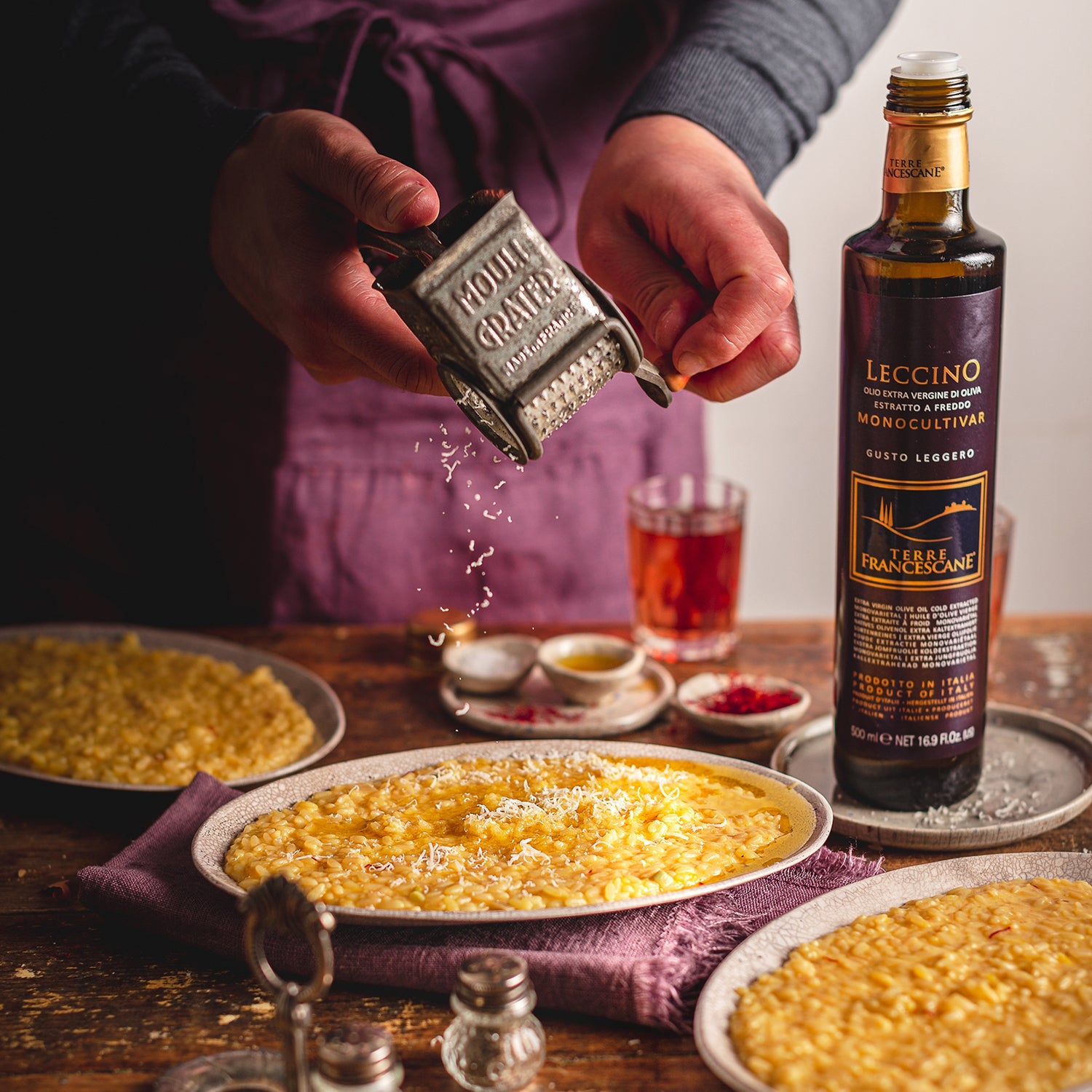Cook grates cheese over risotto beside an open 500 ml bottle of Terre Francescane Leccino olive oil.