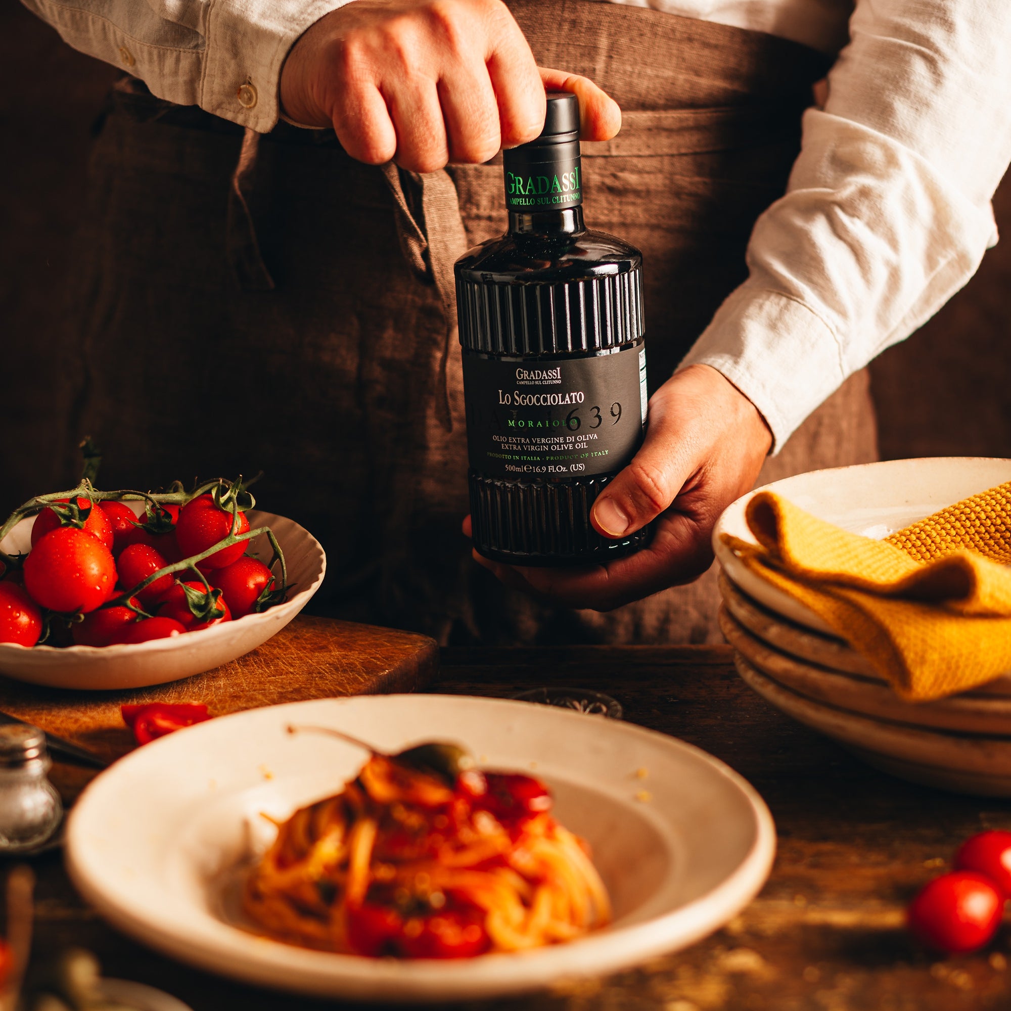 Holding Gradassi Lo Sgocciolato Moraiolo 500 ml bottle near tomato pasta and bowls of fresh tomatoes.