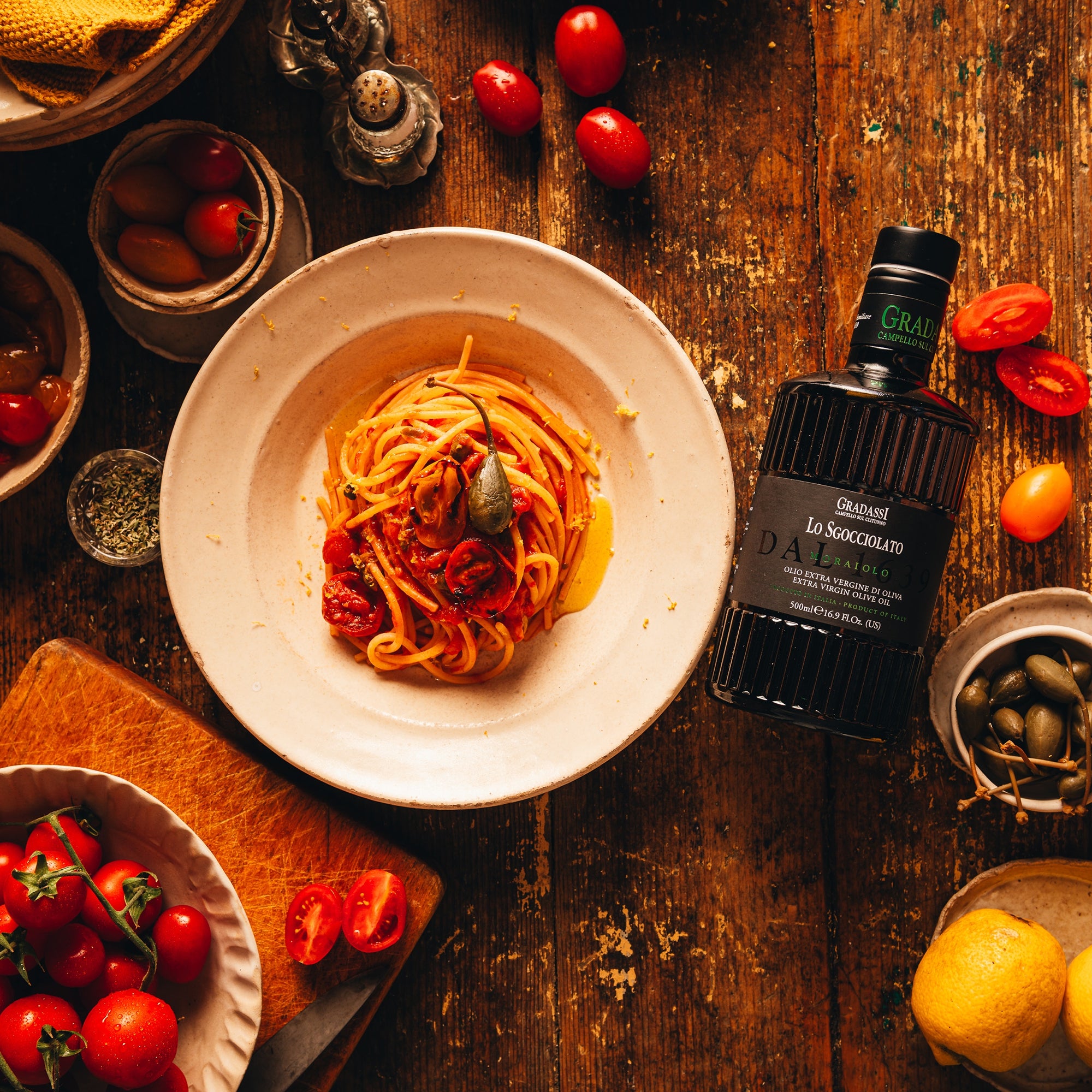 View of Gradassi Lo Sgocciolato Moraiolo 500 ml bottle beside tomato pasta, olives, tomatoes, and lemons.