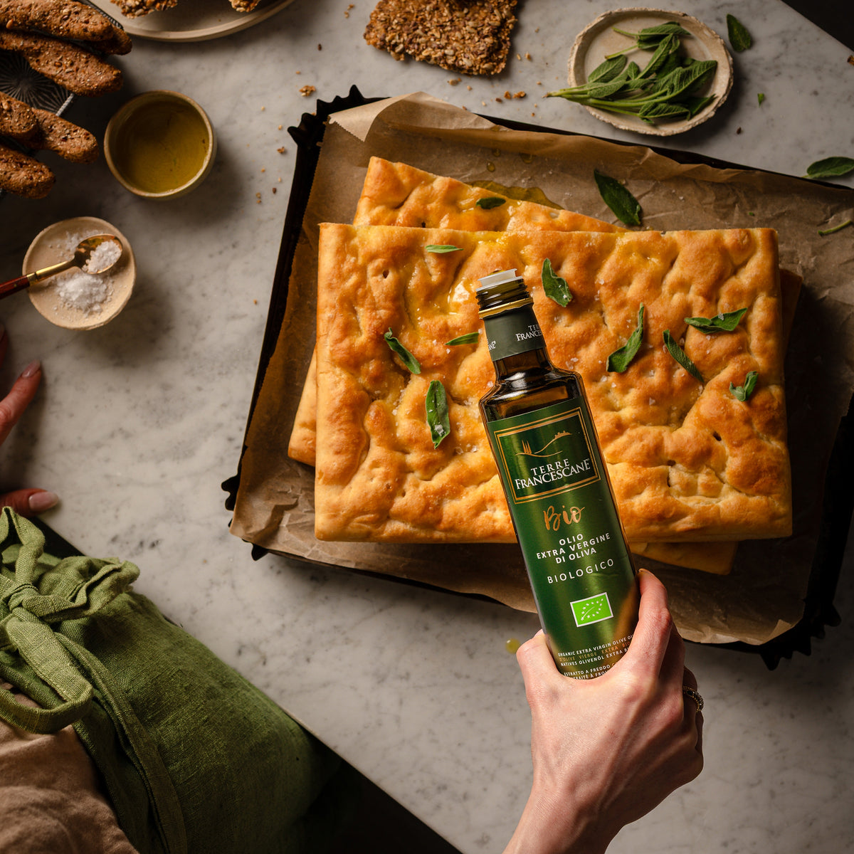 Holding Terre Francescane Organic olive oil bottle over freshly baked focaccia garnished with herbs.