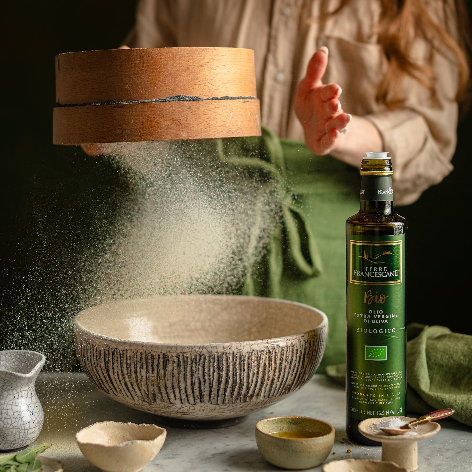 Terre Francescane Organic 500 ml olive oil bottle beside a mixing bowl as flour is sifted for baking.