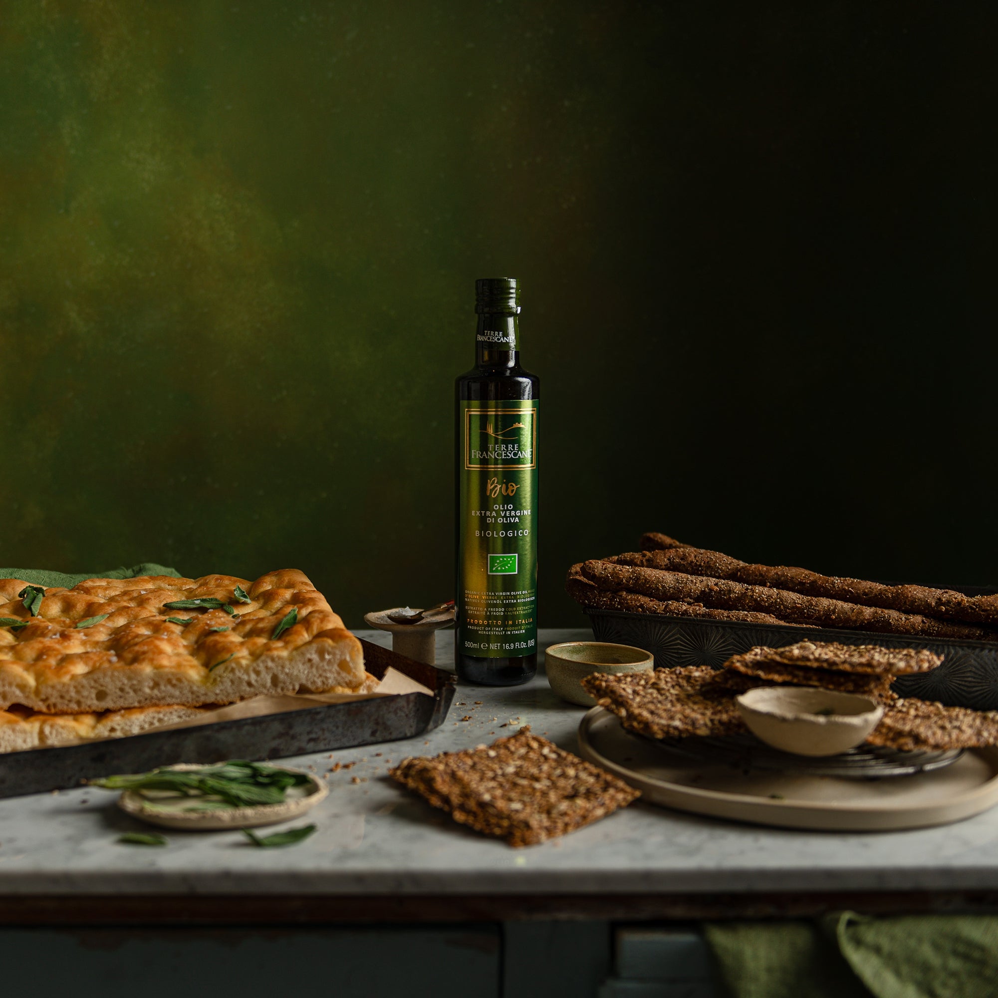 Terre Francescane Organic 500 ml olive oil bottle beside focaccia, seeded crackers, breadsticks, and herbs.