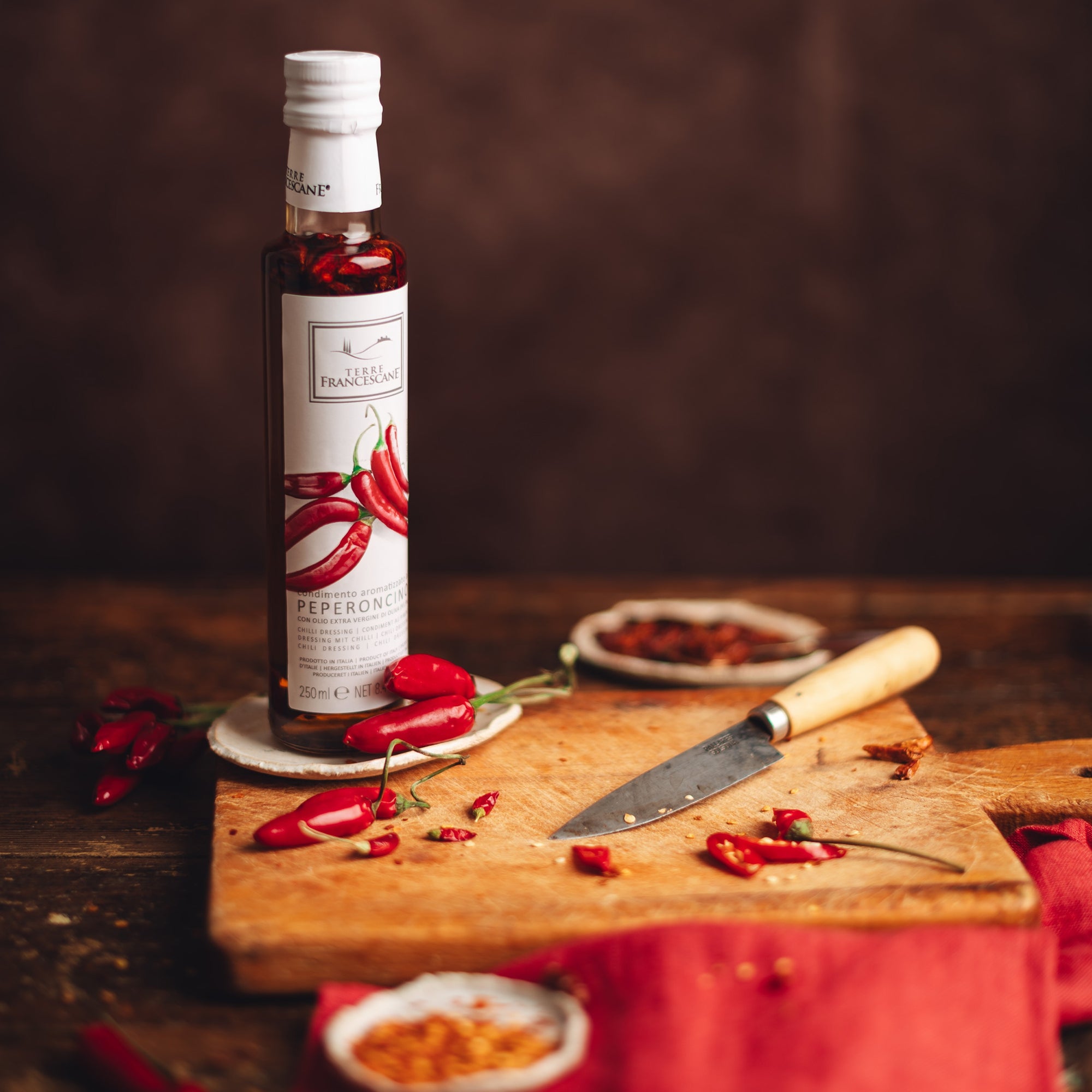 250 ml Terre Francescane Peperoncino bottle beside fresh chilies, cutting board, and dried chili flakes.