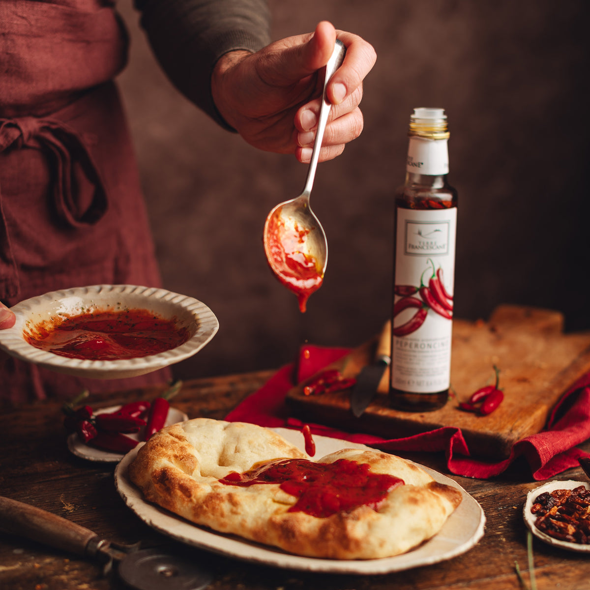 Spooning seasoned tomato sauce onto bread, beside Terre Francescane Peperoncino 250 ml bottle.