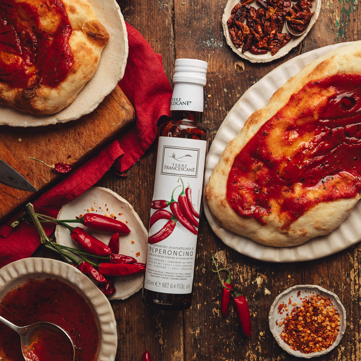 Terre Francescane Peperoncino 250 ml bottle surrounded by chili peppers, tomato-topped bread, sauce, and chili flakes.