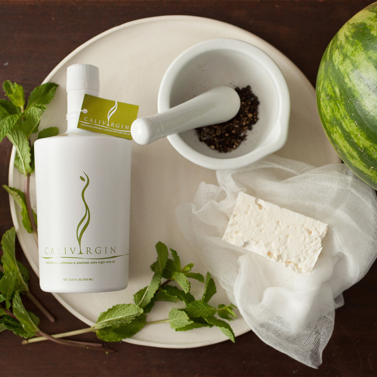 Calivirgin Premium 500 ml bottle with fresh mint, feta cheese, peppercorns, and a watermelon.