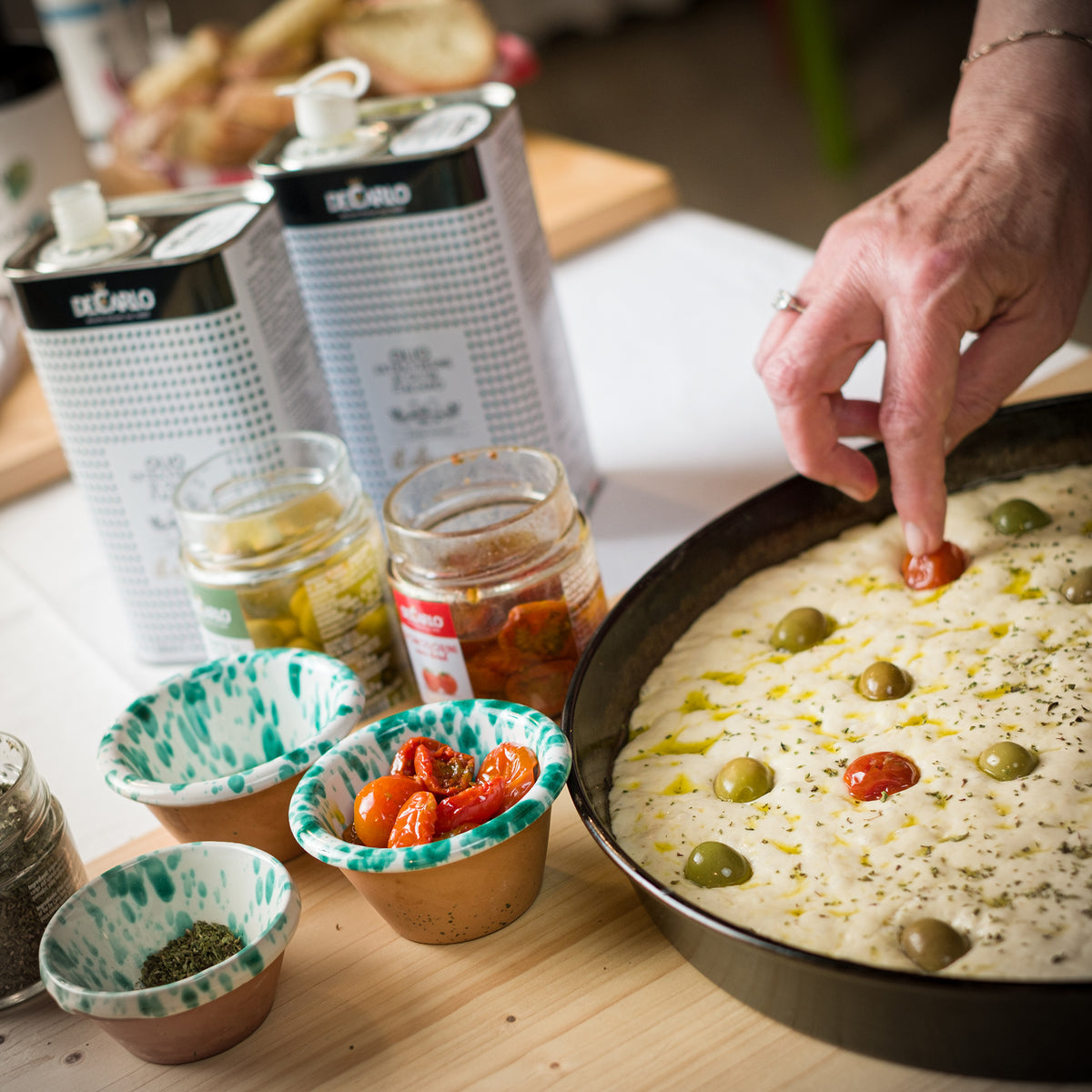De Carlo il Classico tins beside focaccia being topped with De Carlo Bella di Cerignola Olives, and De Carlo Semi-Dried Cherry Tomatoes.