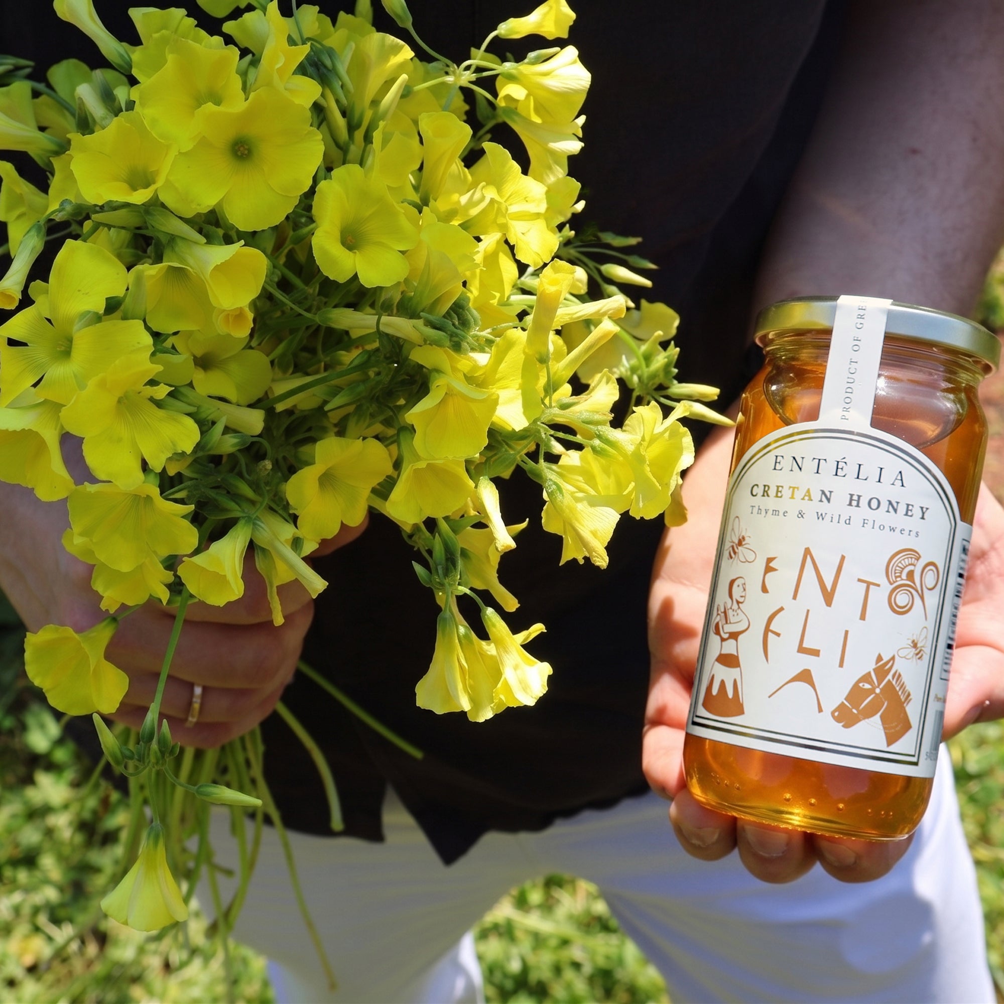 Holding a 450g jar of Entelia Thyme & Wild Flowers Cretan Honey, and a bouquet of yellow wildflowers.