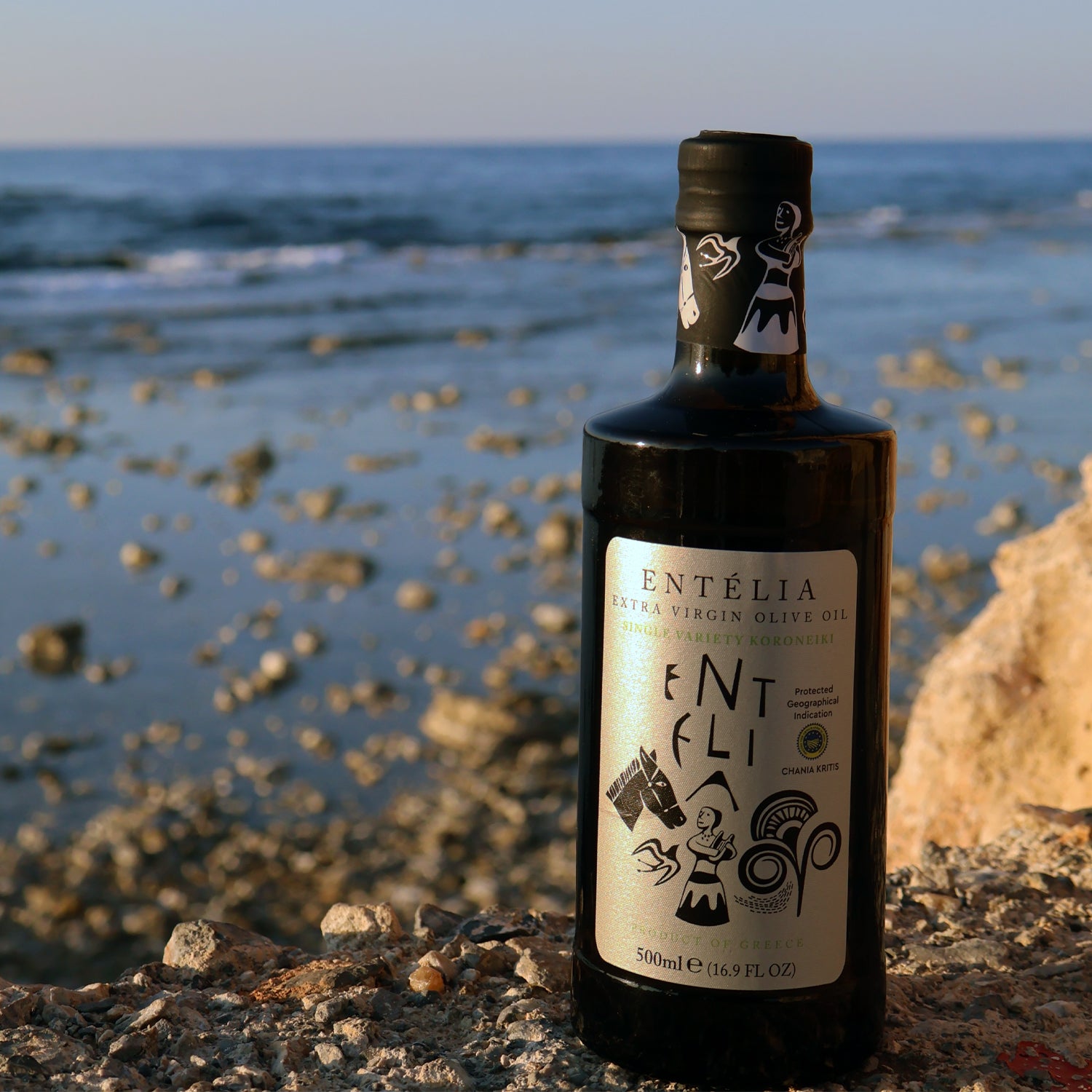 Entelia extra virgin olive oil bottle beside a rocky seashore.