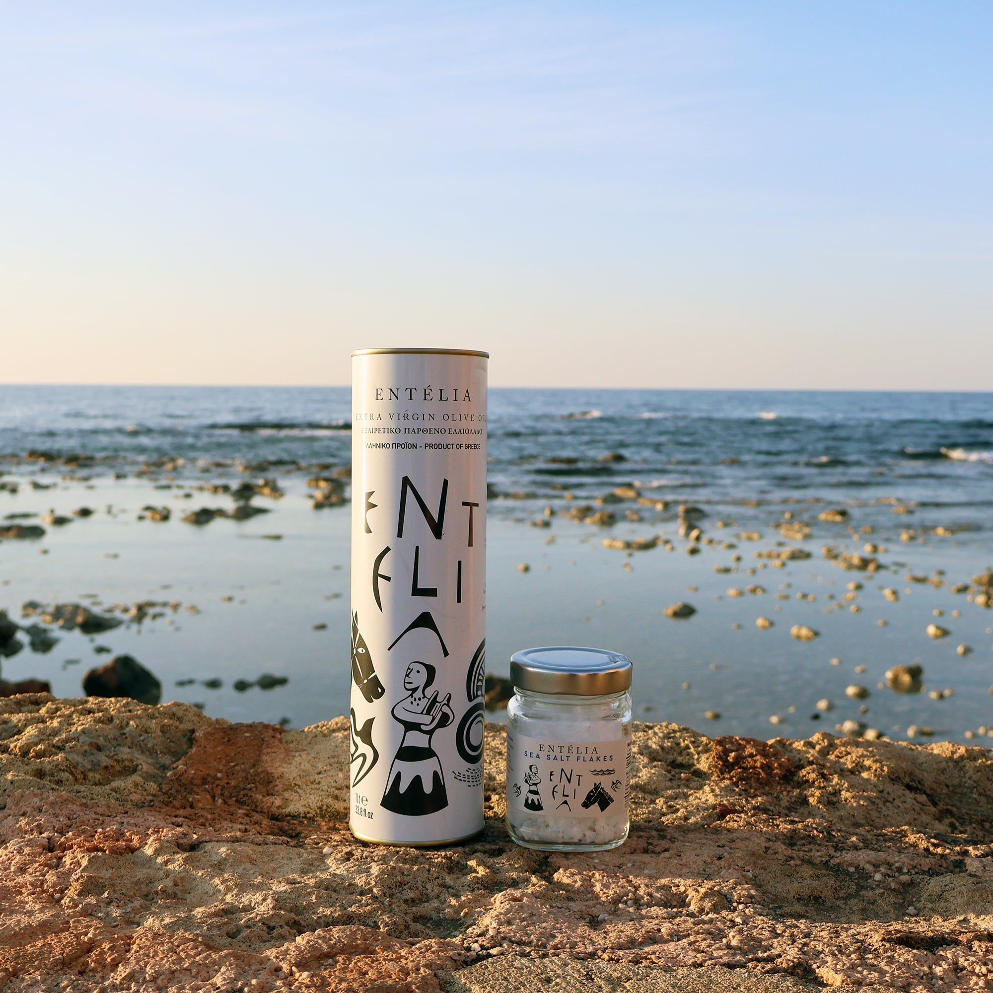 An Entelia 1L extra virgin olive oil can, and an Entelia Pyramid Sea Salt jar on coastal rocks beside the sea.