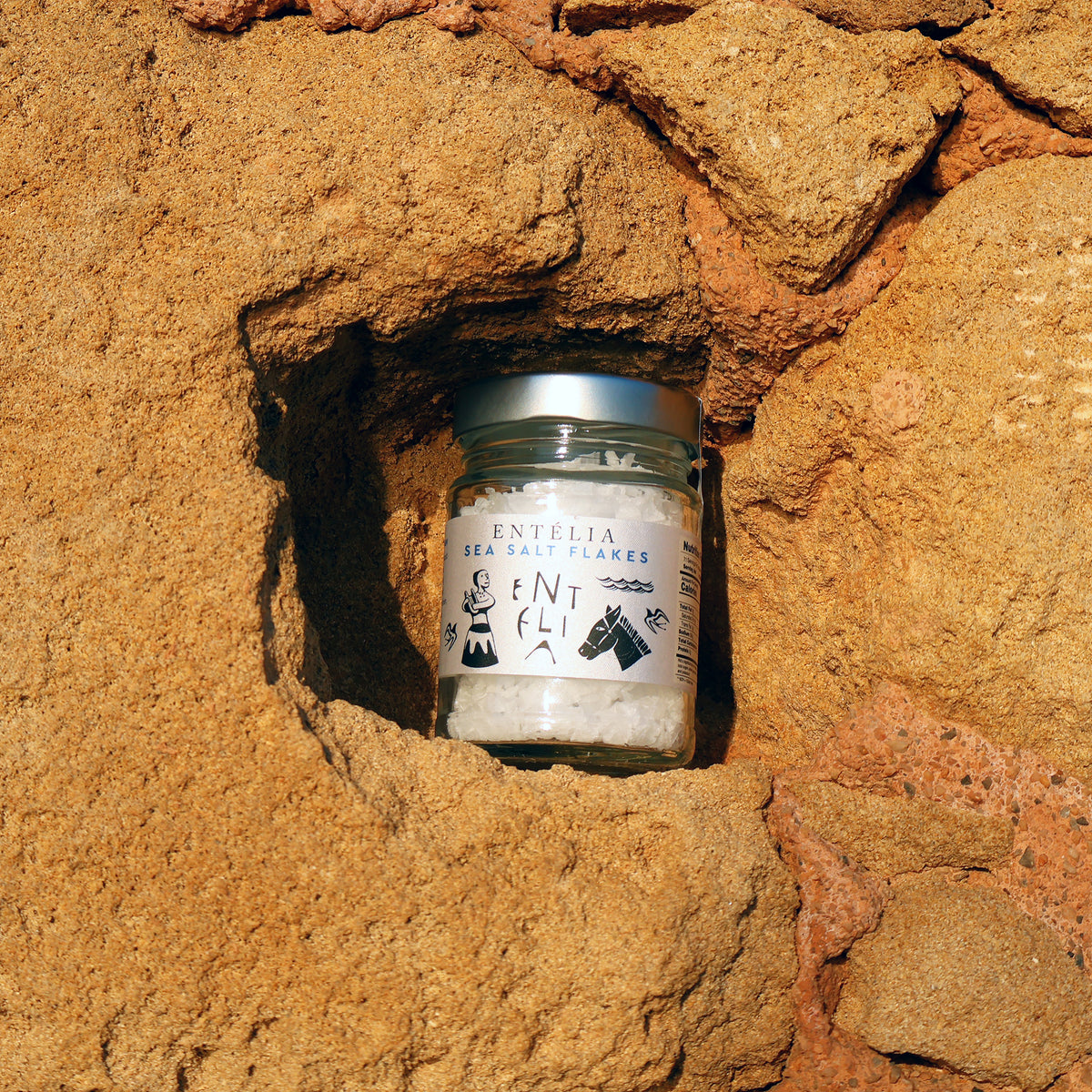 Jar of Entelia Pyramid Sea Salt Flakes nestled within coastal rocks.