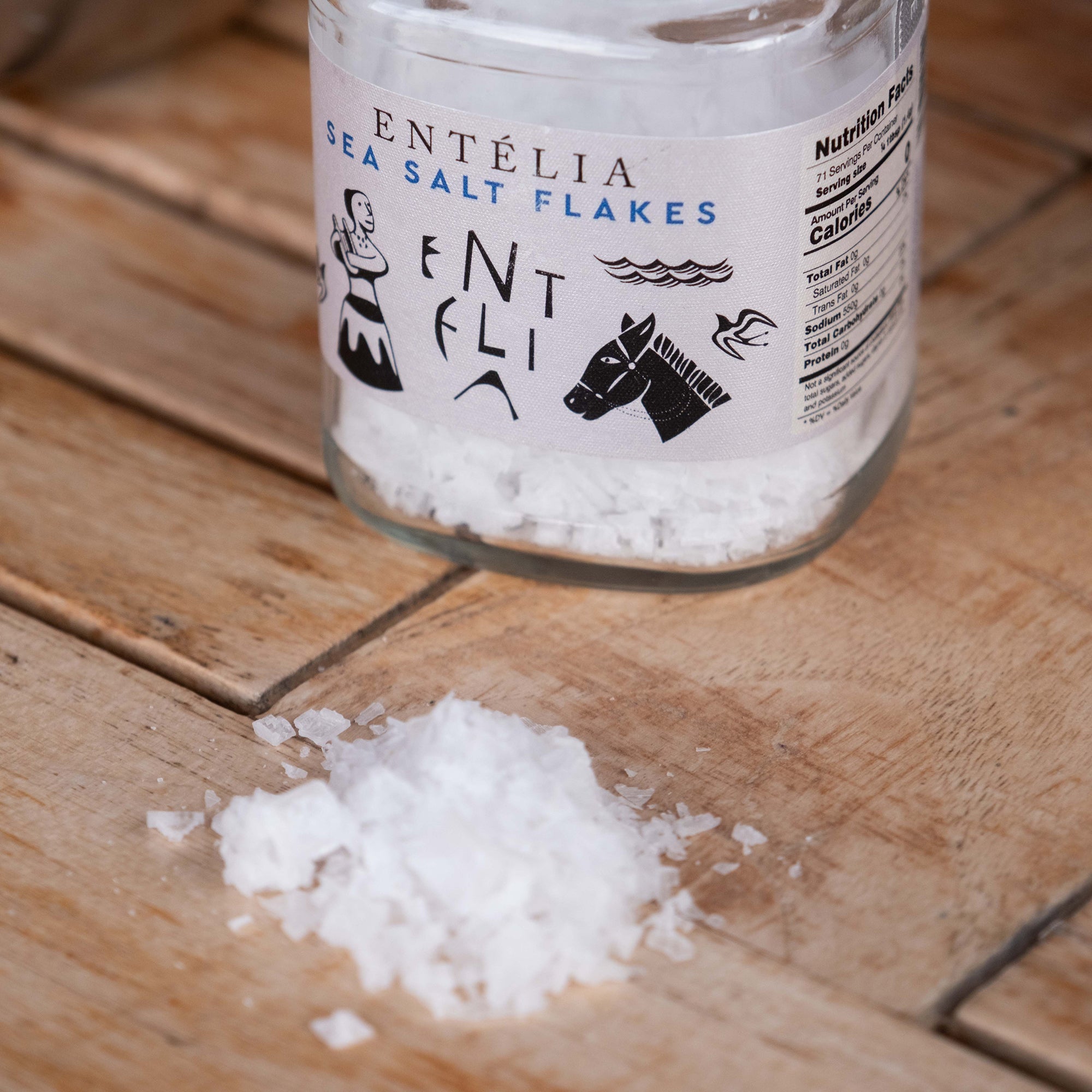 Close-up of Entelia Pyramid Sea Salt Flakes jar with loose flakes scattered across a wooden table.