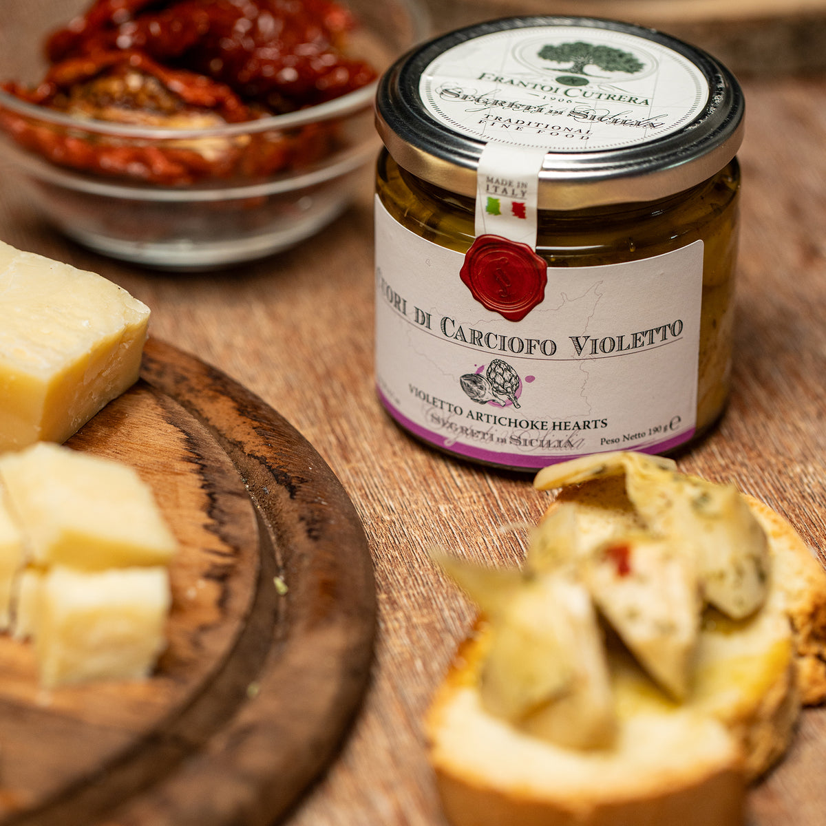 Frantoi Cutrera Violetto Artichoke Hearts jar beside cheese, bread, and sun-dried tomatoes.