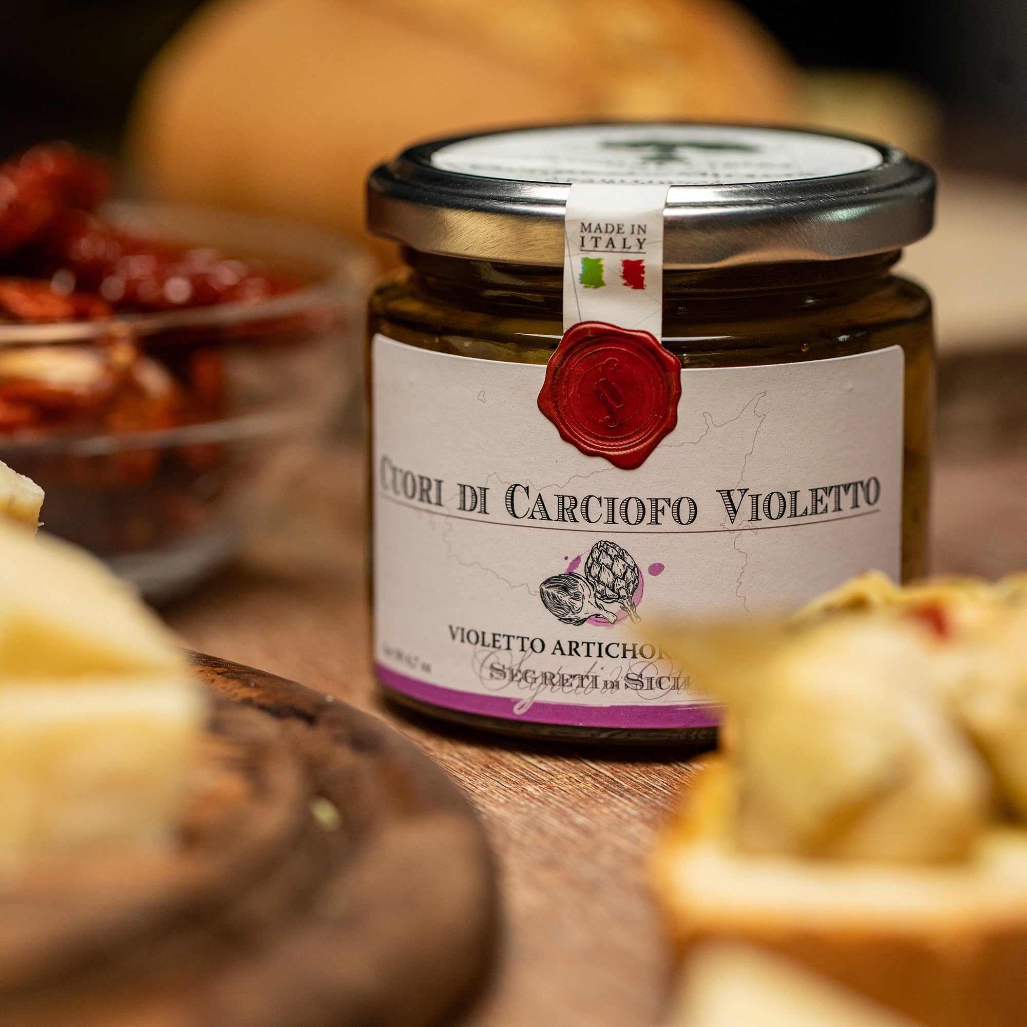 Close-up of Frantoi Cutrera Violetto Artichoke Hearts jar surrounded by bread, cheese, and sun-dried tomatoes.